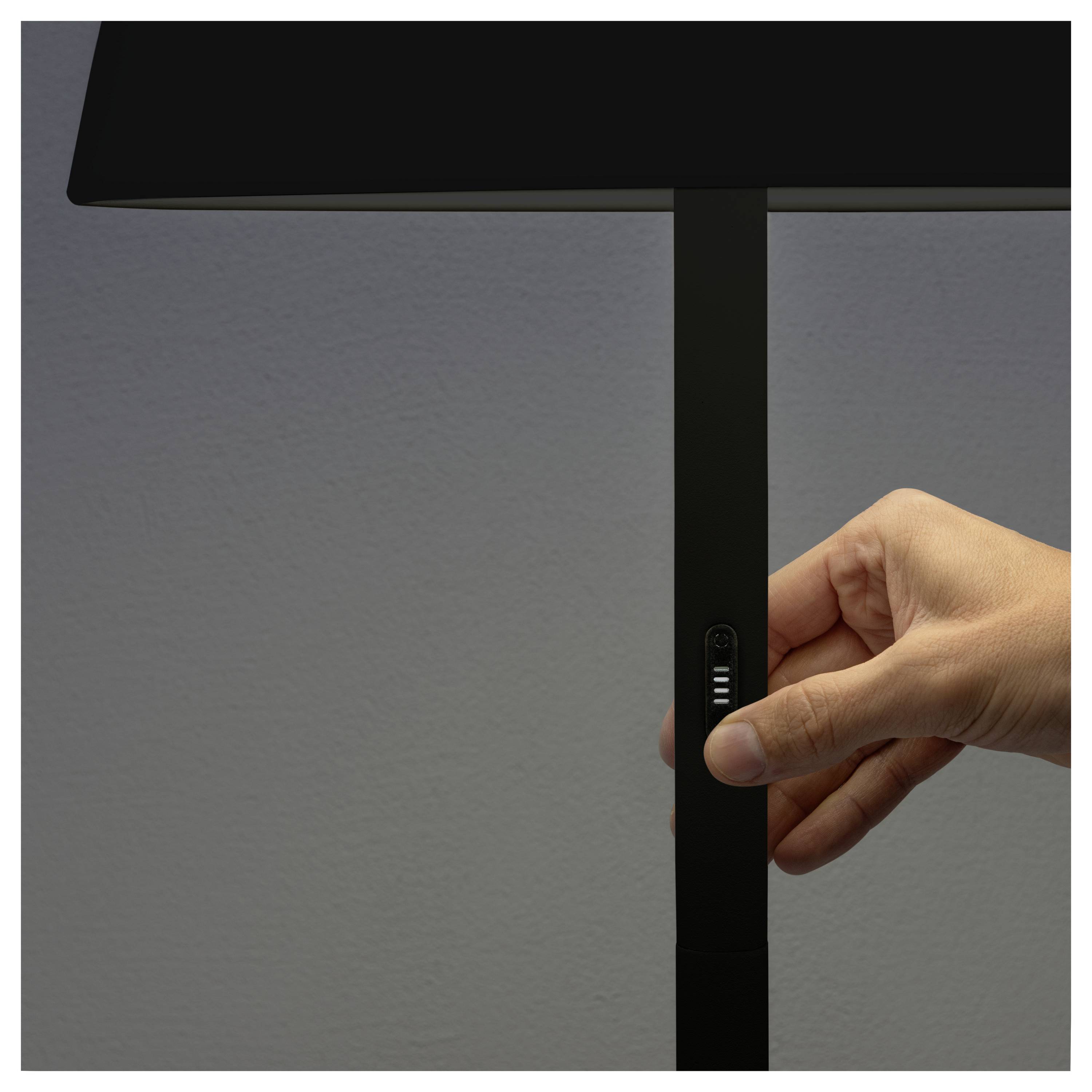 Een hand die de dimmerknop bedient op een moderne zwarte lamp, tegen een grijze muur op de achtergrond.