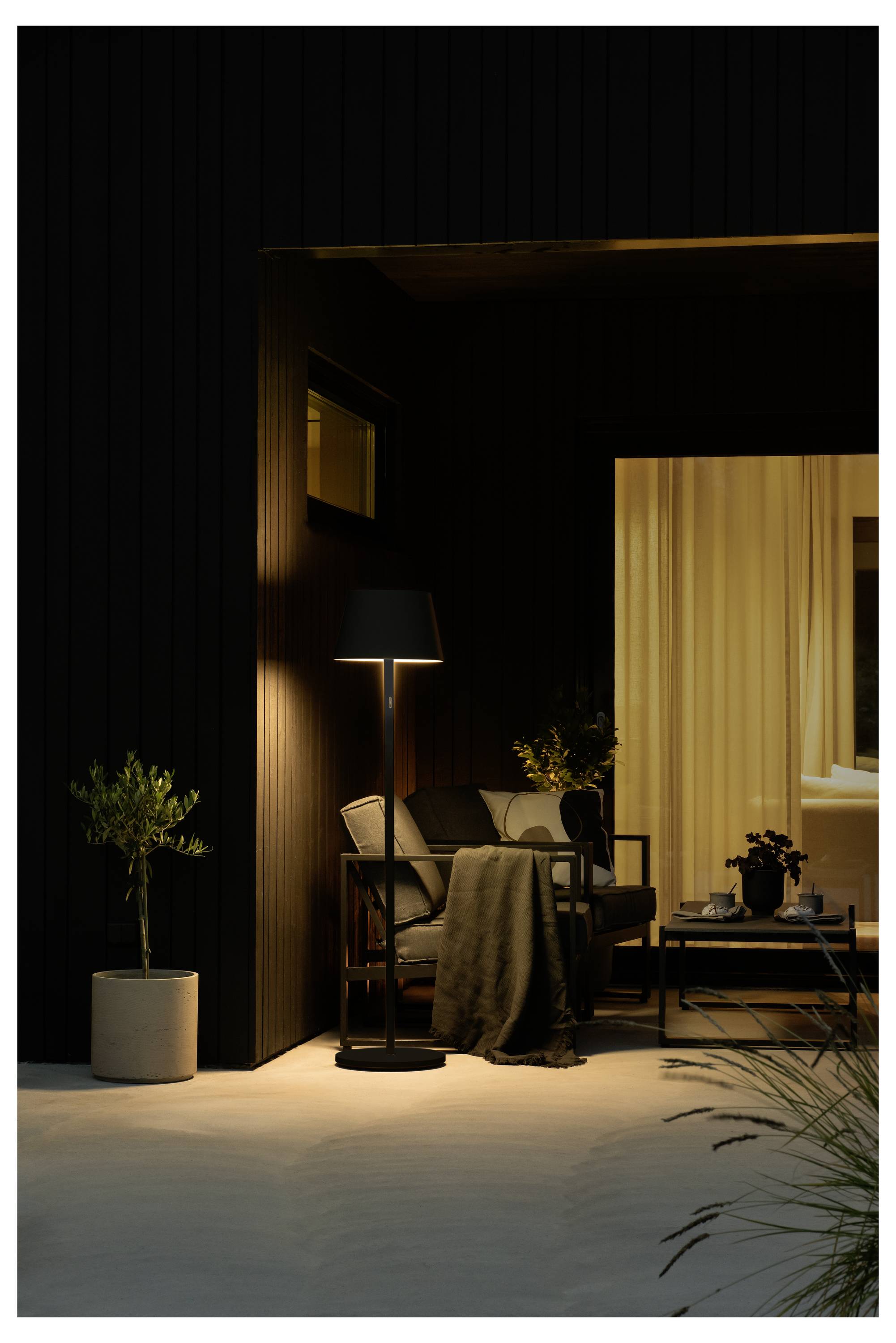 Buitenzitruimte met een lamp, bank en planten. Het warme licht van binnen contrasteert met de donkere buitenkant, waardoor een gezellige sfeer ontstaat.