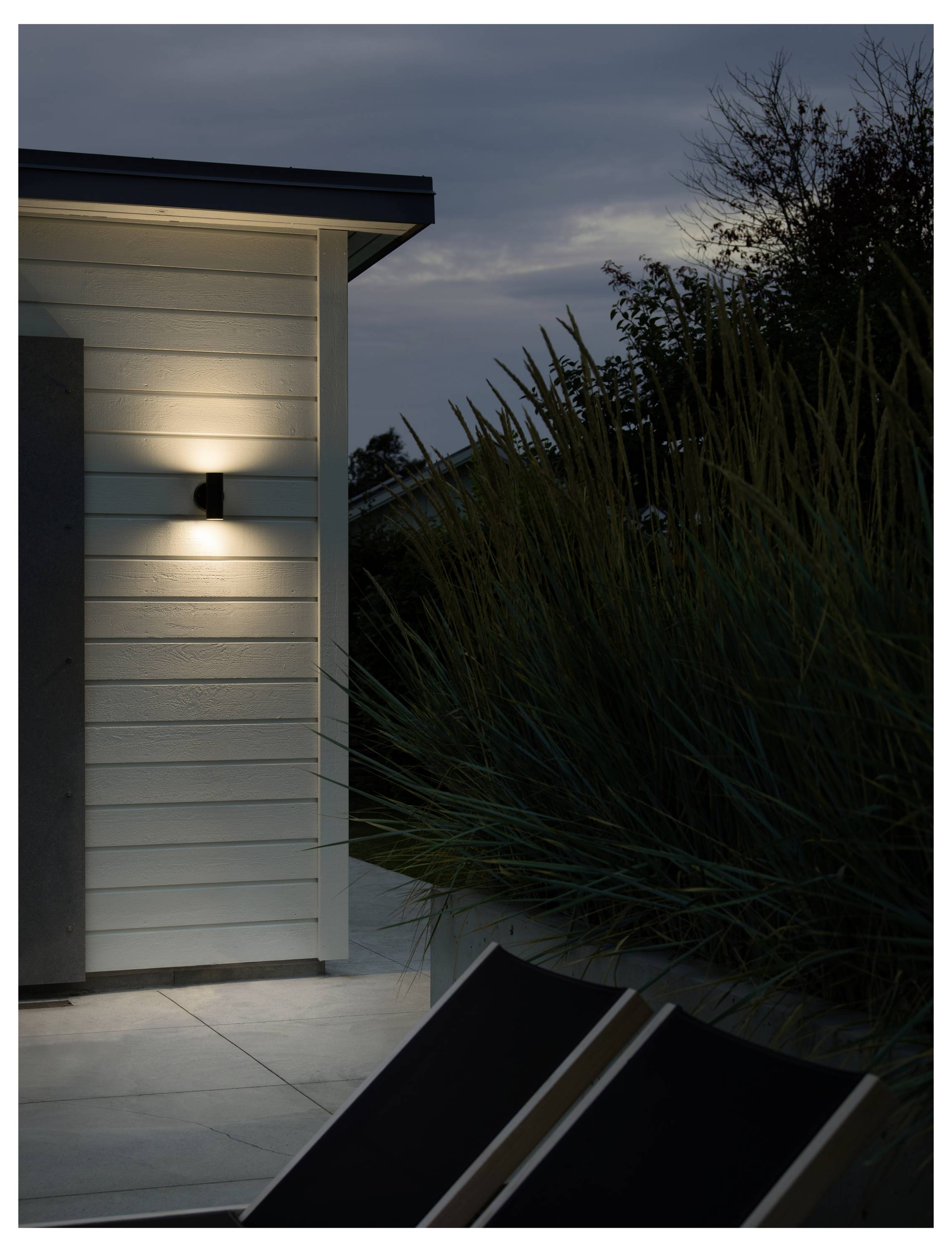 Een moderne buitenwandlamp verlicht de buitenkant van een huis bij schemering, met hoge grassen en een terrasstoel in de buurt, waardoor een gezellige sfeer ontstaat.