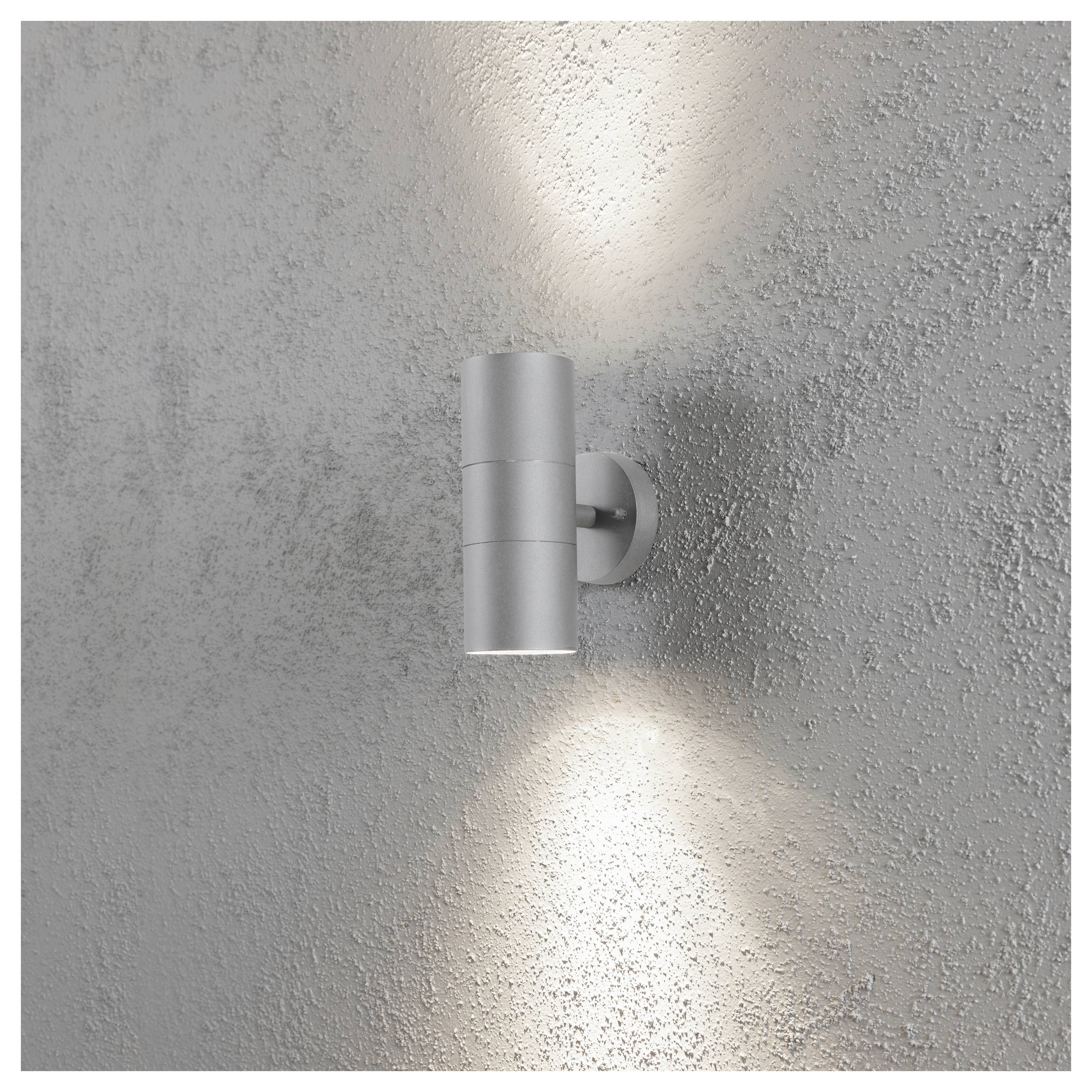 Een cilindrische zilveren wandlamp gemonteerd op een gestructureerde grijze muur, die lichtbundels naar boven en beneden werpt voor sfeerverlichting.