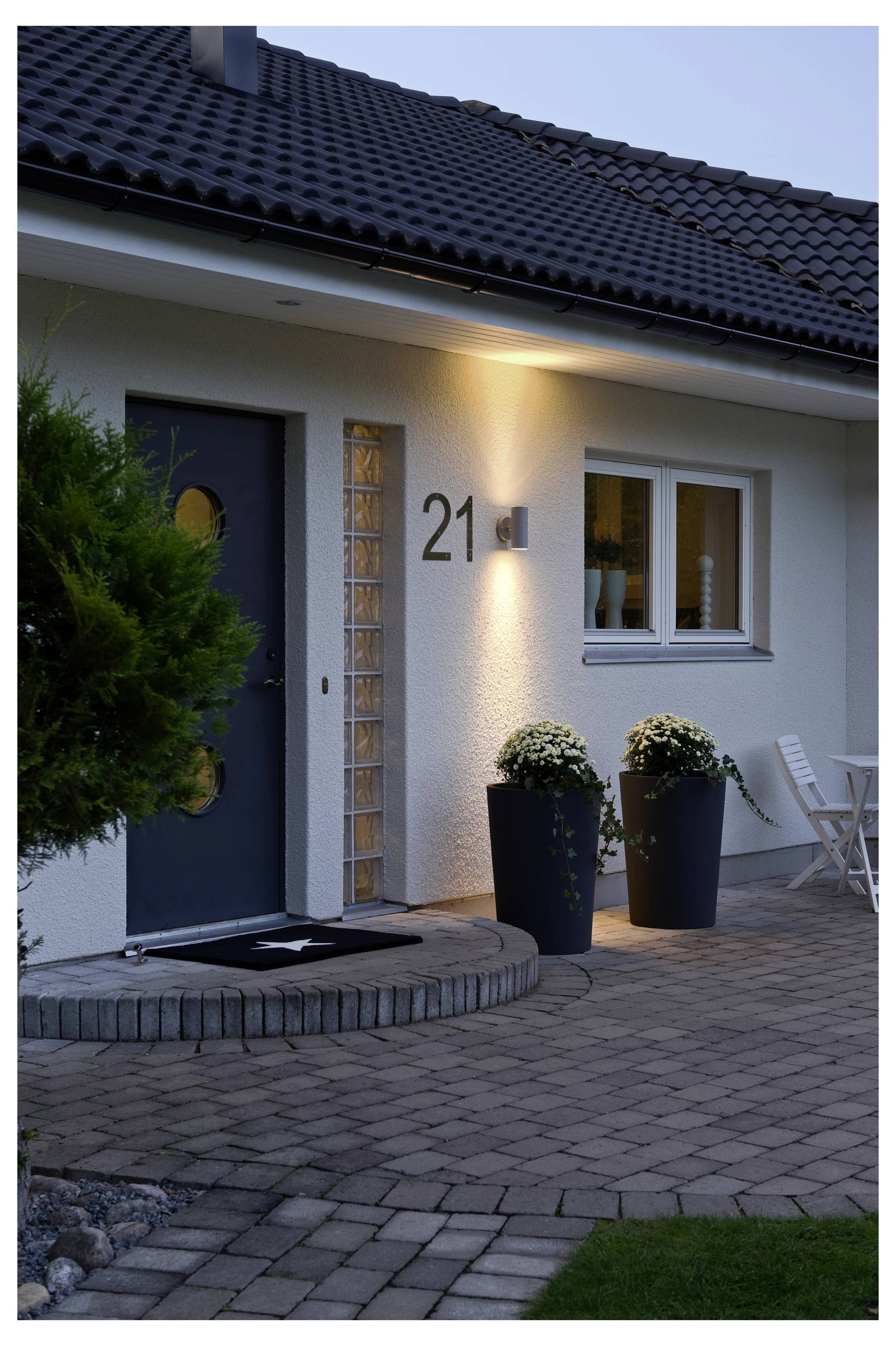 De afbeelding toont de entree van een modern huis, met een donkere deur met ronde ramen, decoratieve planten en het nummer '21' op de muur.