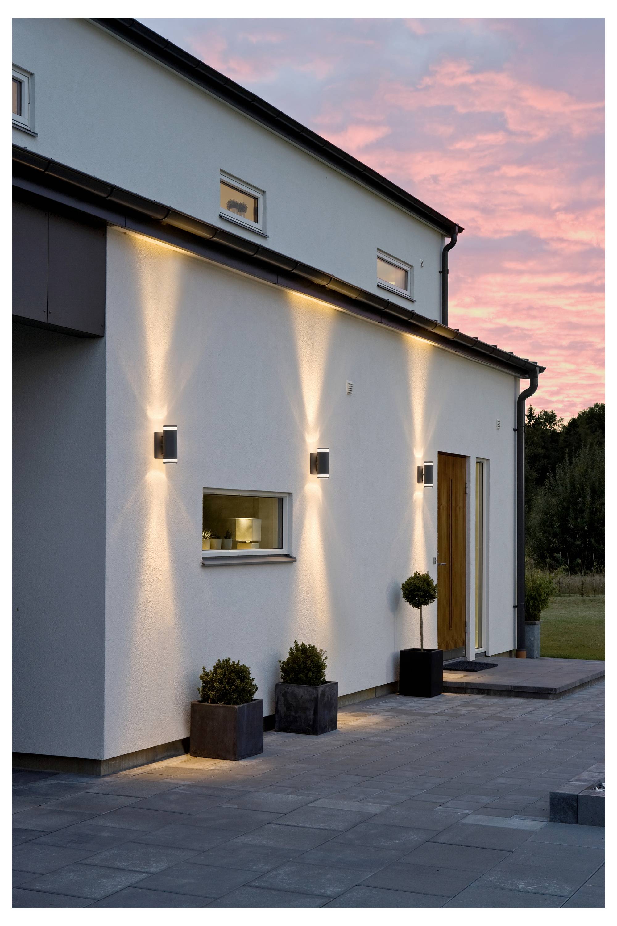 Modern huis exterieur met verlichte wandlampen bij het invallen van de avond, met een houten deur, kleine ramen en potplanten op het terras.