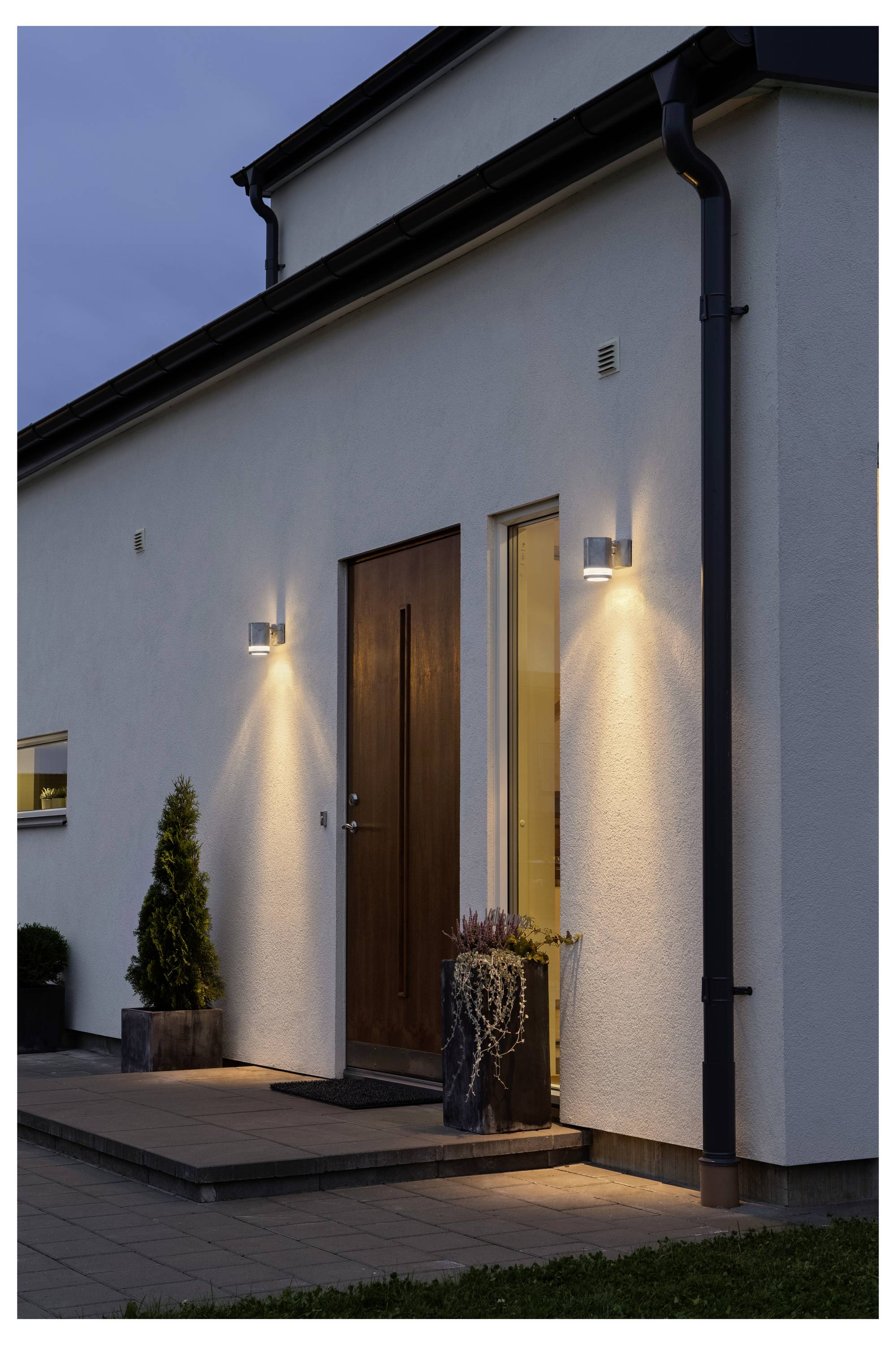 Een moderne huisingang bij het invallen van de avond met aan de muur bevestigde verlichting die de houten deur verlicht en een kleine potplant naast de deuropening.