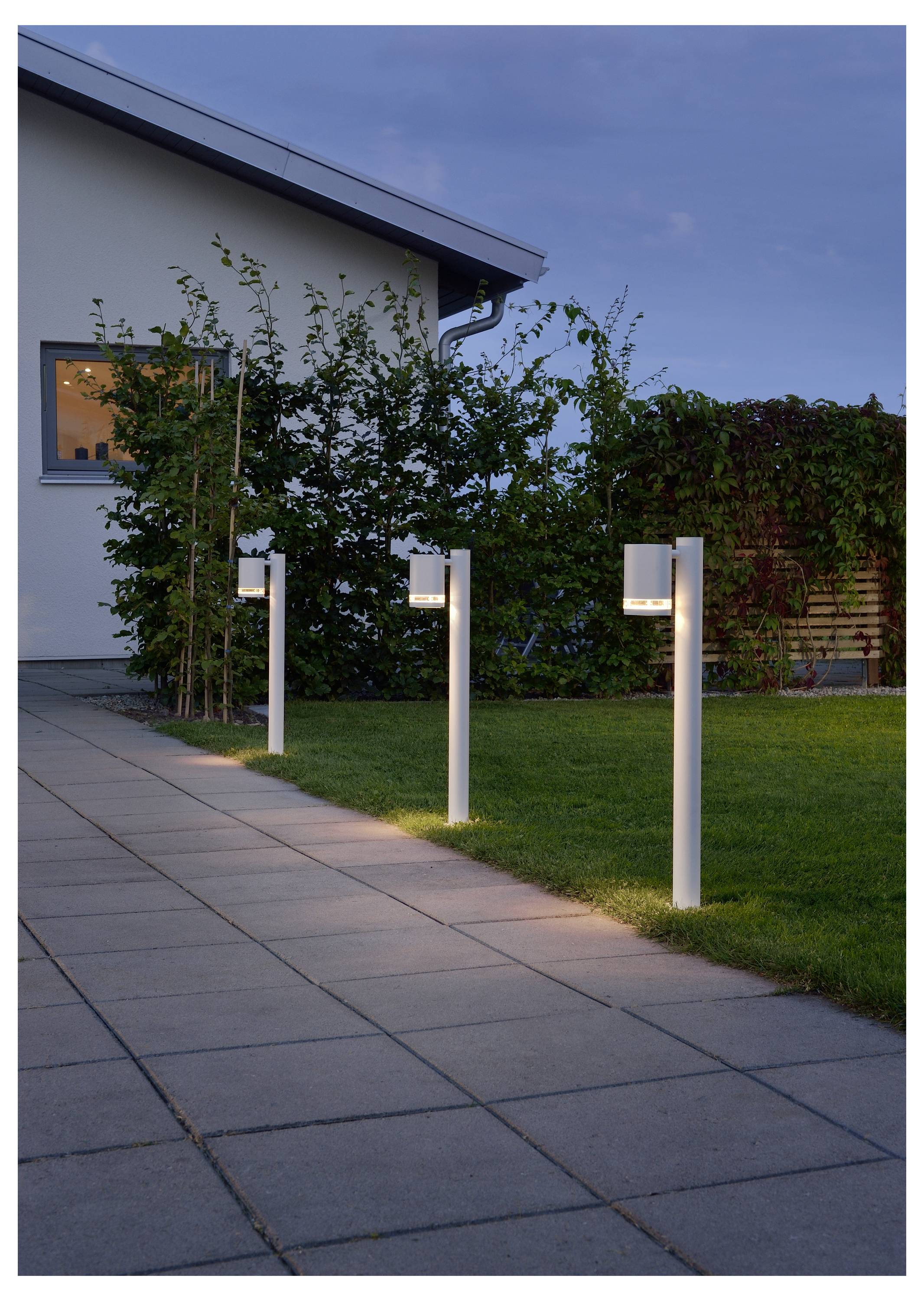 Drie moderne buitenlampen verlichten een tuinpad naast een huis bij het invallen van de avond.
