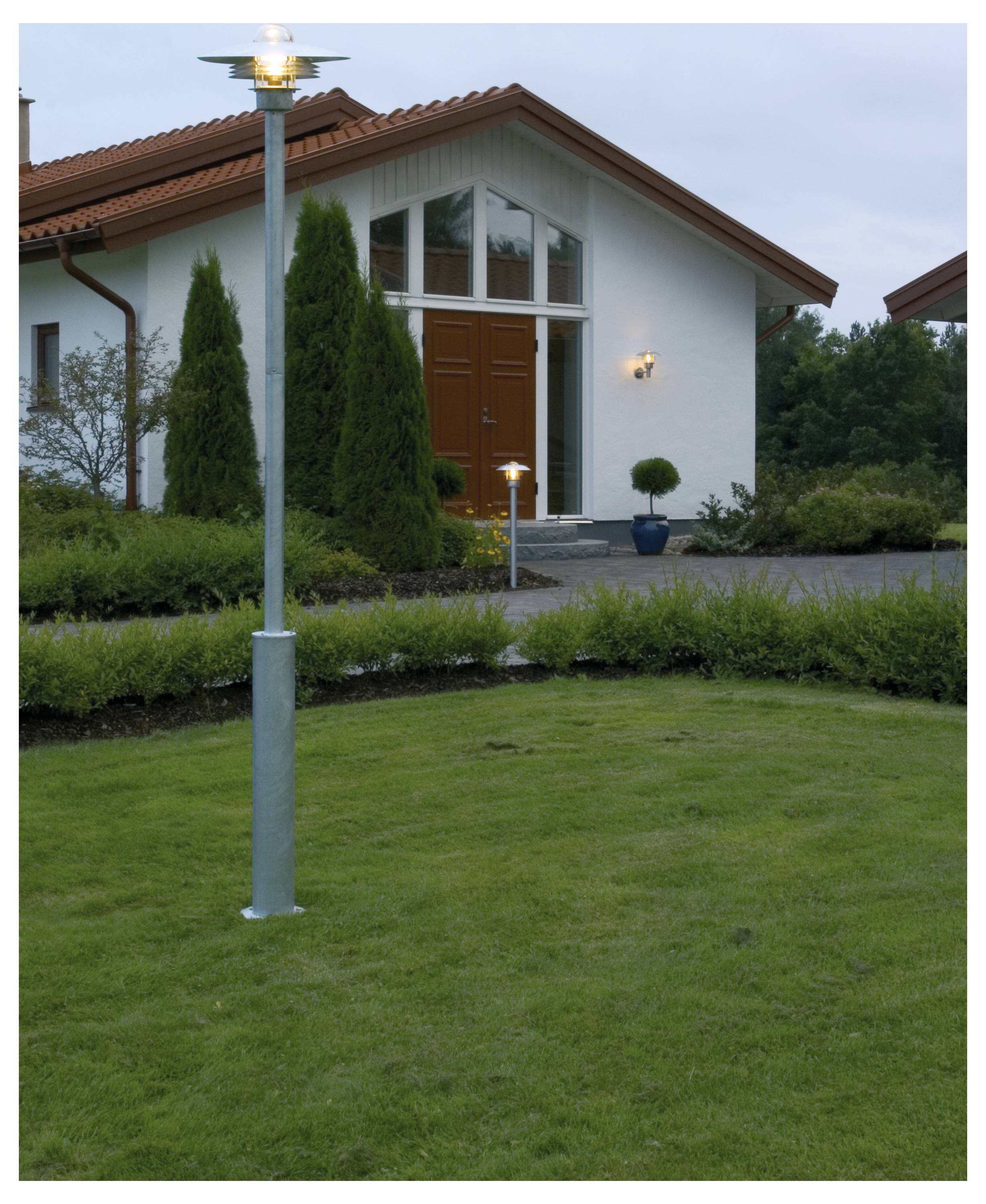 Een modern huis met een rood dak en een grote houten voordeur wordt omringd door een goed onderhouden tuin, met een verlichte lantaarnpaal.