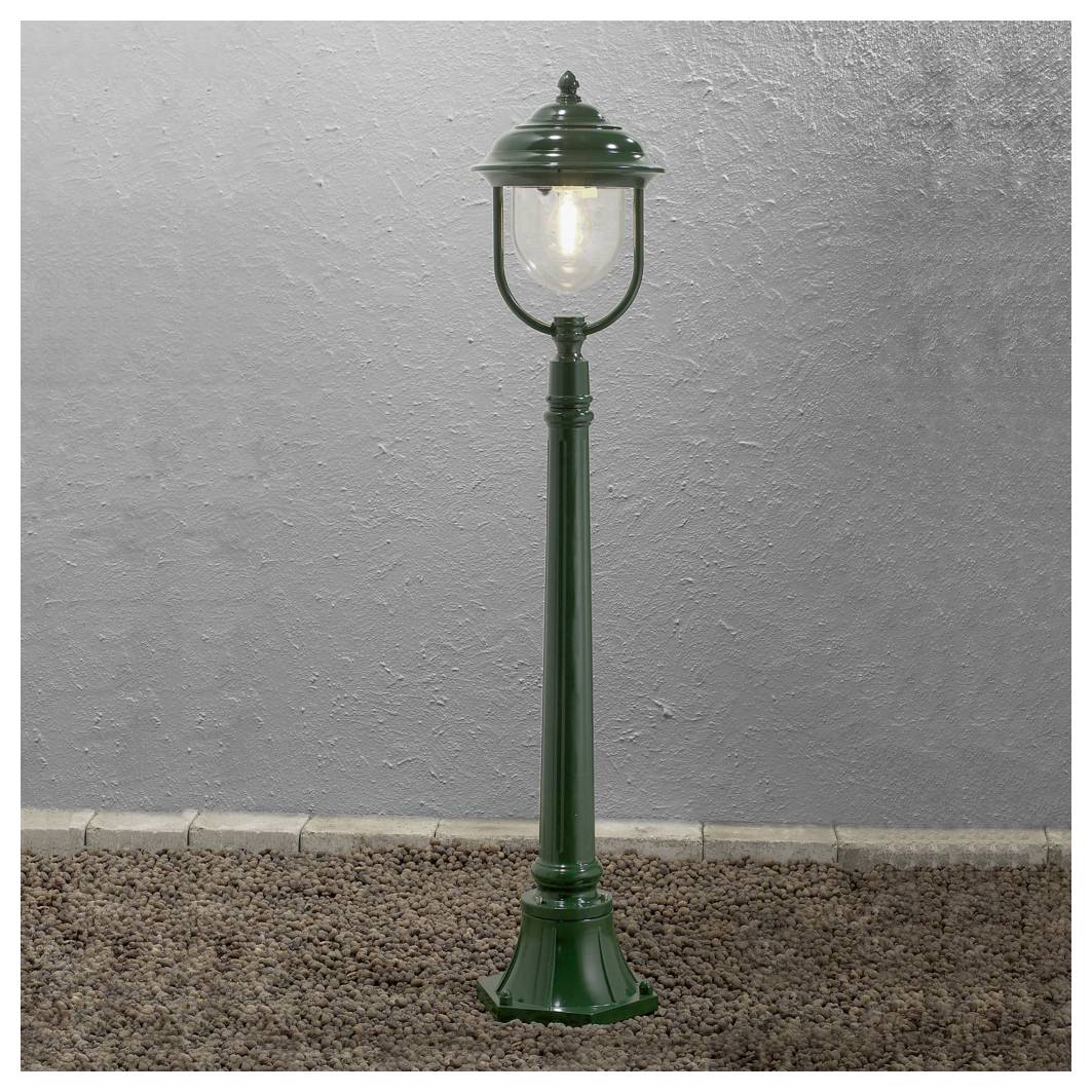 Een groene vintage-stijl lantaarnpaal staat op een grindpad tegen een gestructureerde grijze muur, waarbij hij zijn omgeving verlicht.