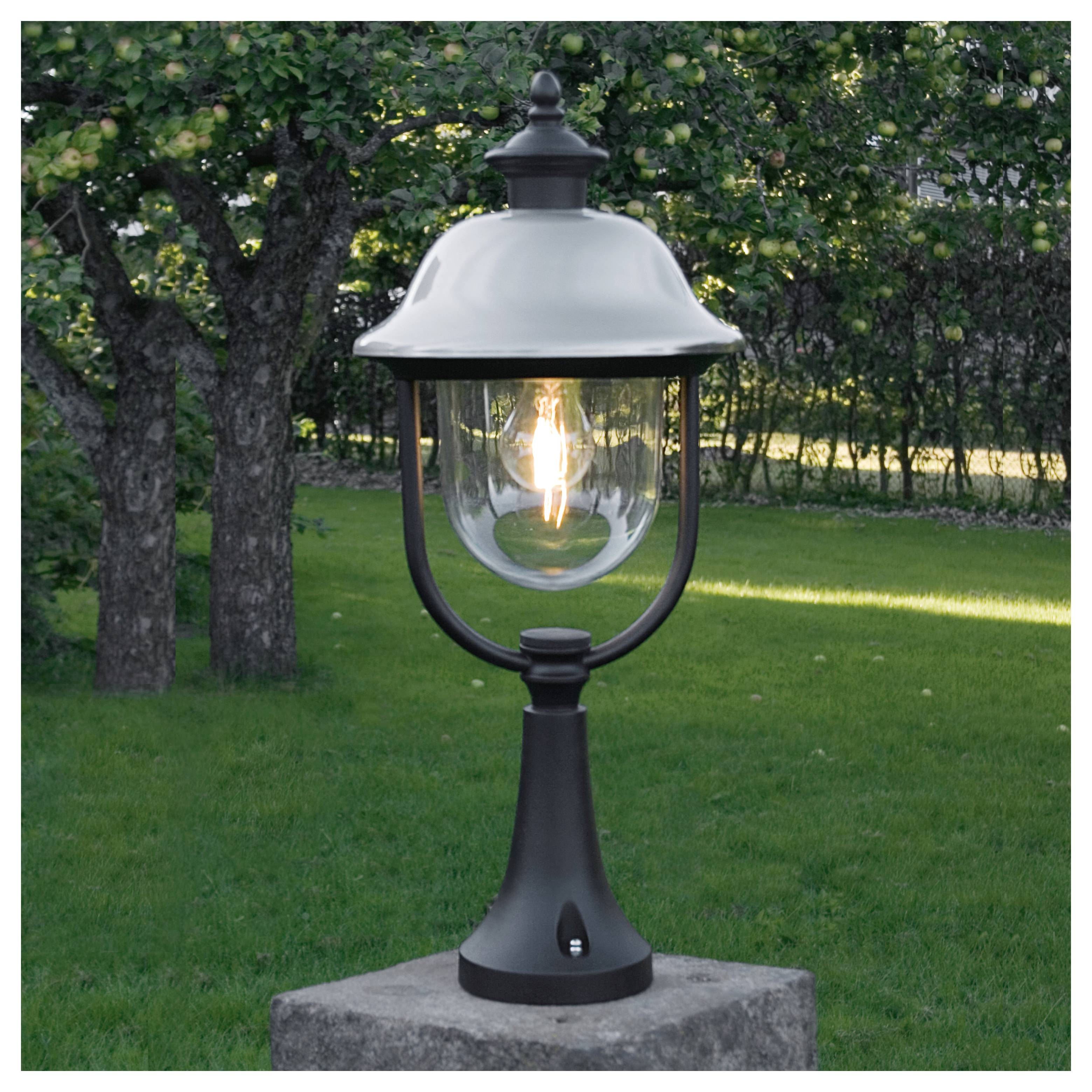 Een buitenlantaarn met een gloeiende lamp is bevestigd op een paal in een groen gazongebied, omringd door bomen.