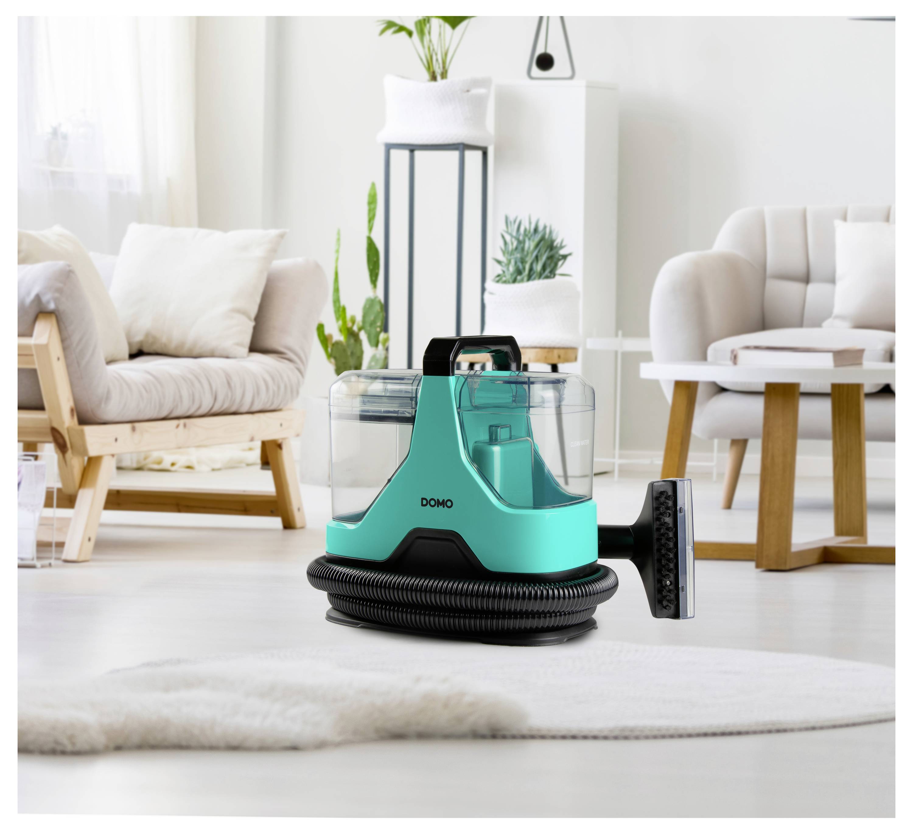 Een turquoise stofzuiger met een slang staat op een wit tapijt in een moderne, lichte woonkamer met een stoel, plant en kussens.