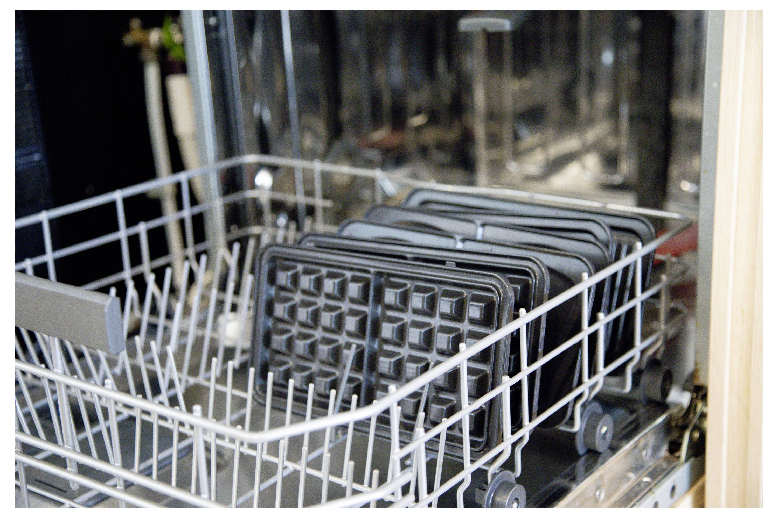 Een openstaande vaatwasser met een metalen rekje dat verschillende zwarte platen van een wafelijzer vasthoudt, klaar om schoongemaakt te worden.