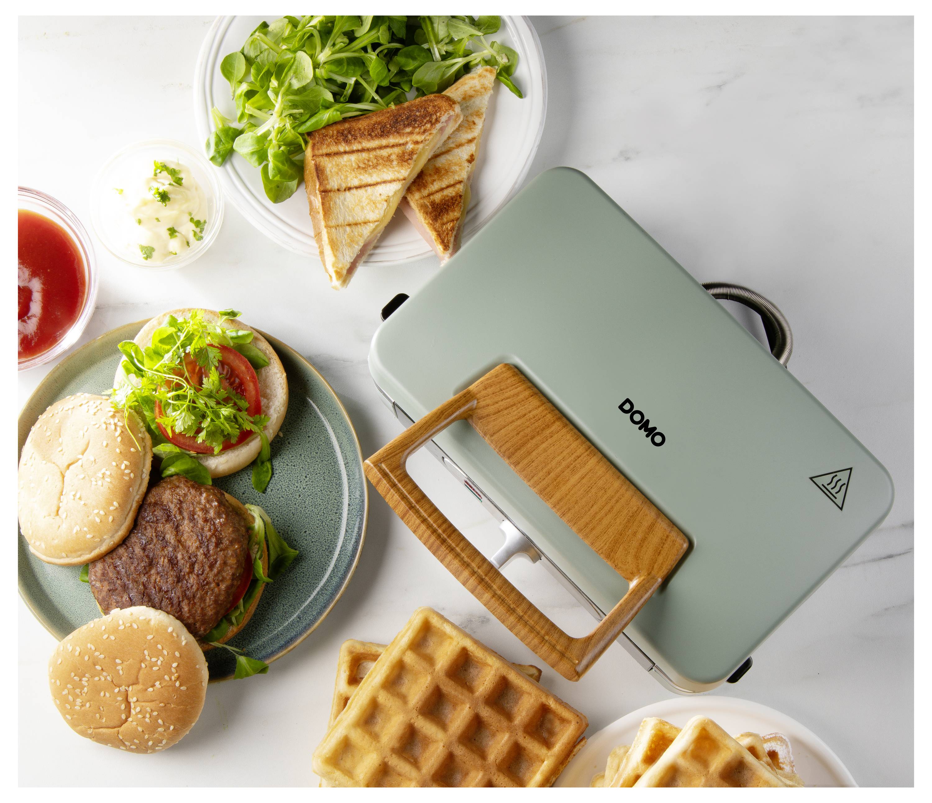 Een groene tosti-ijzer met een houten handvat is omringd door een bord met hamburgerpatties, broodjes, wafels, gegrilde tosti's en salade.