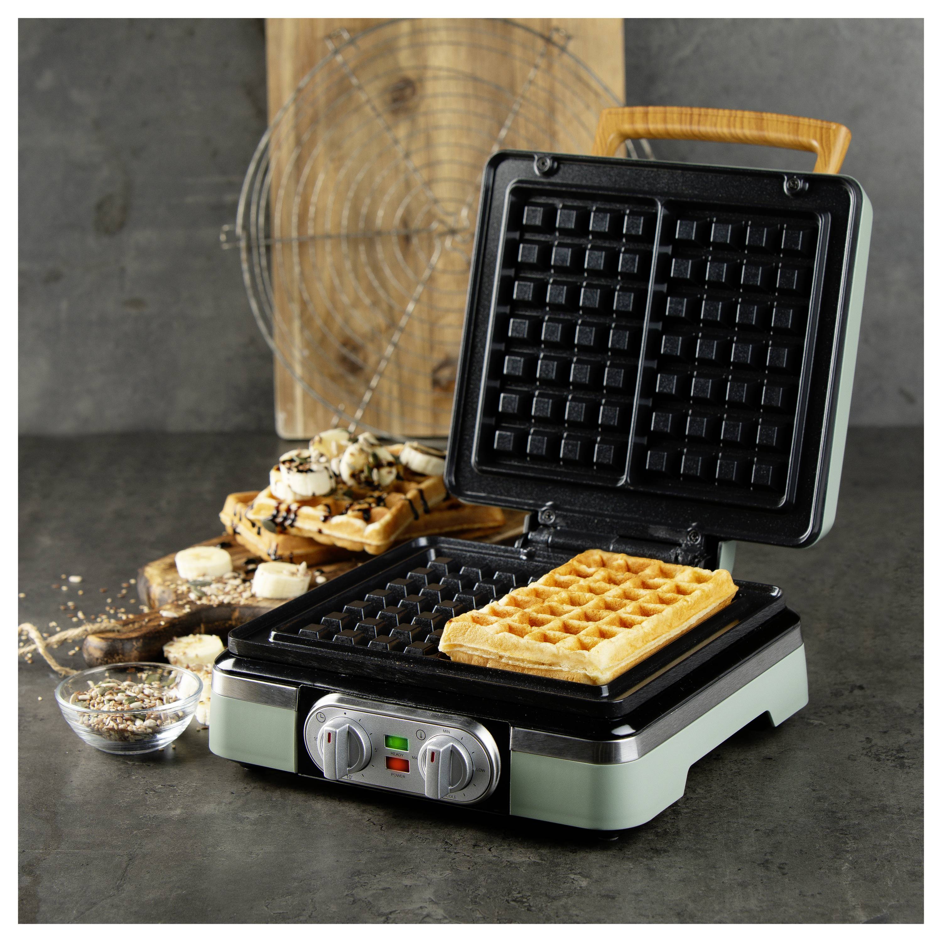Een wafelijzer met een vers gebakken wafel erin, naast een houten plank met gestapelde wafels en een kom met toppings.