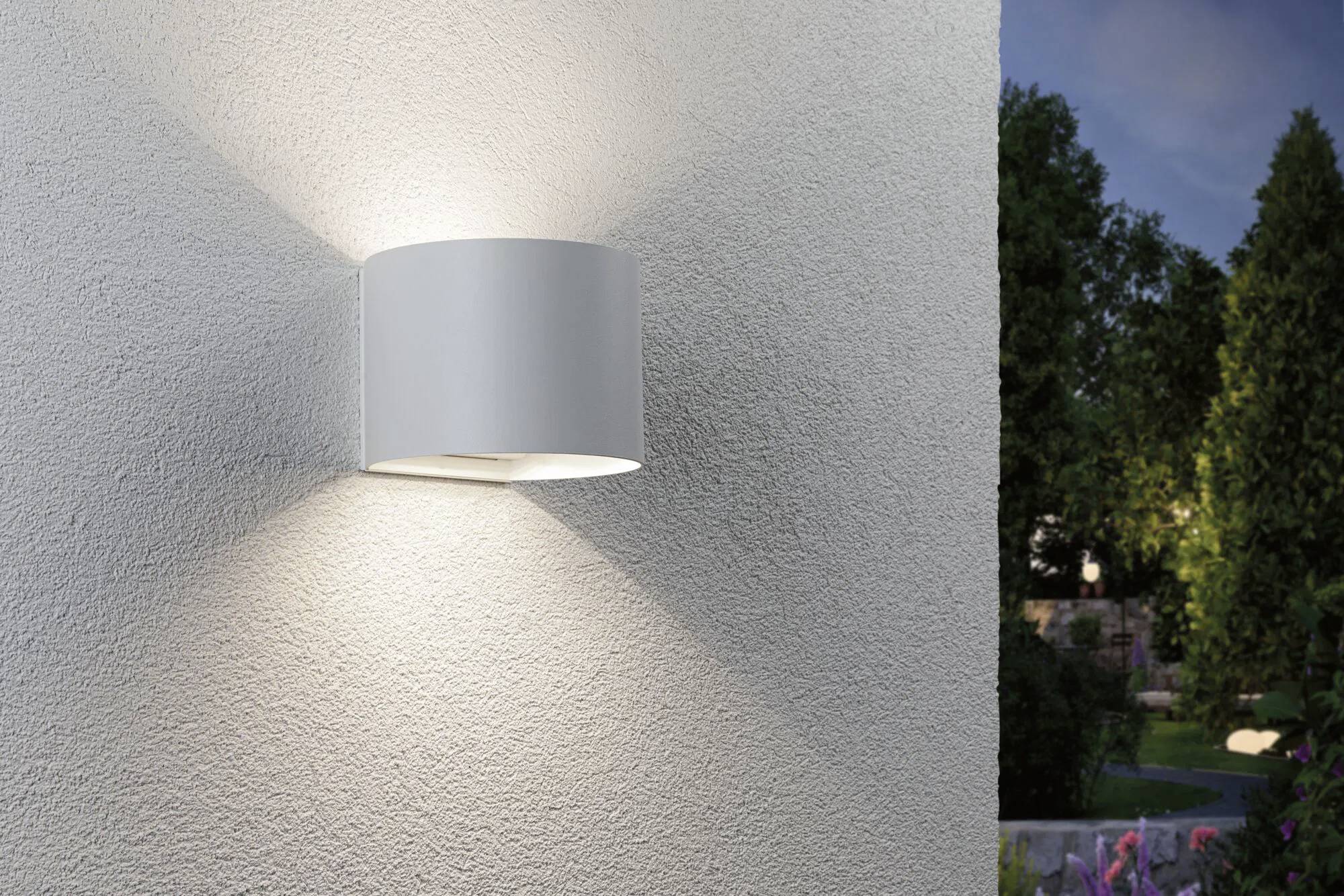 Een moderne buitenwandlamp schijnt tegen een gestructureerde muur, waardoor een sfeervolle avondsfeer ontstaat in een tuinomgeving met bomen.