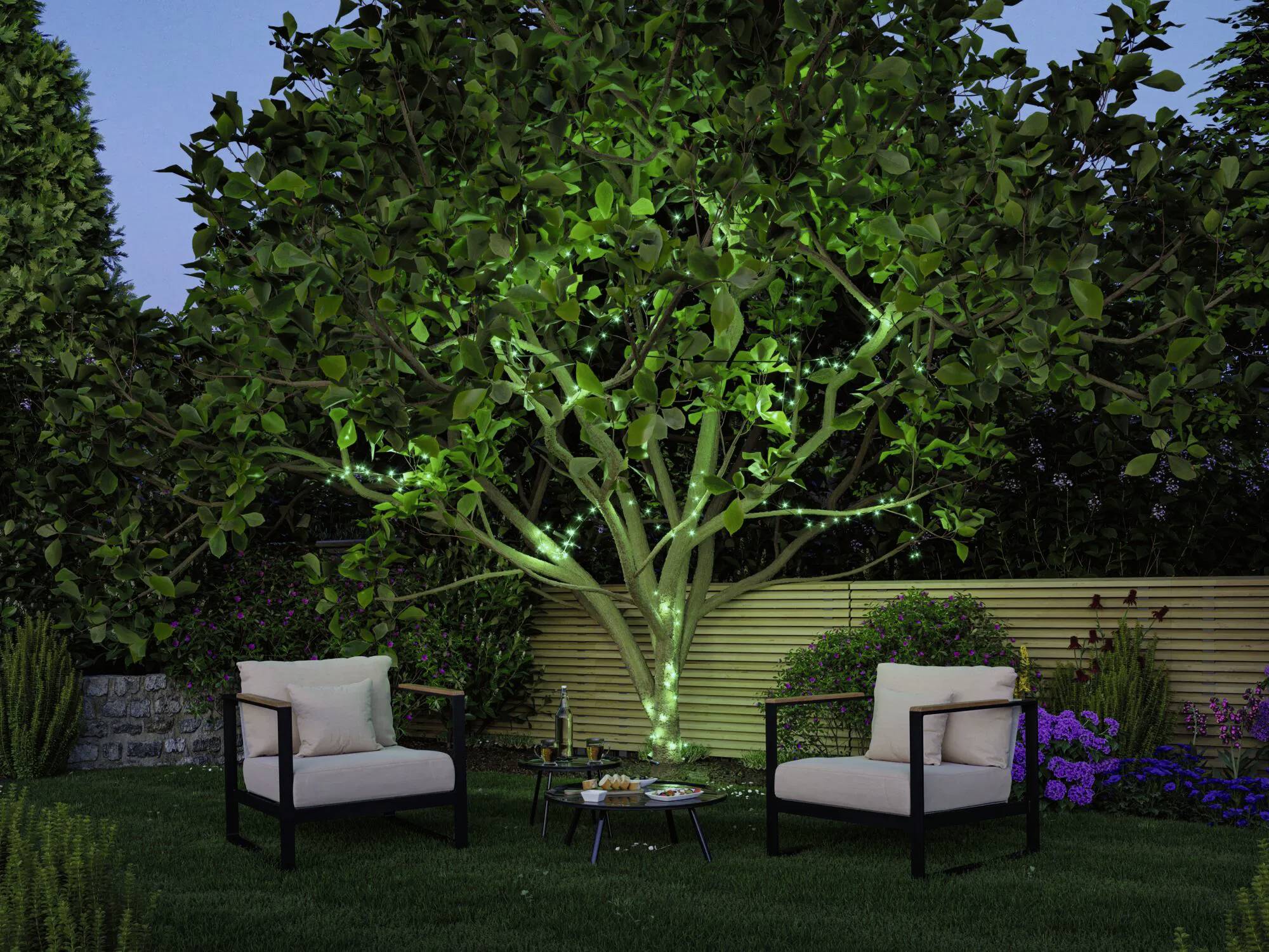 Tuinscène bij het invallen van de avond met een boom die wordt verlicht door groene verlichting. Twee beige stoelen staan tegenover elkaar bij een kleine tafel met hapjes en een kaars.