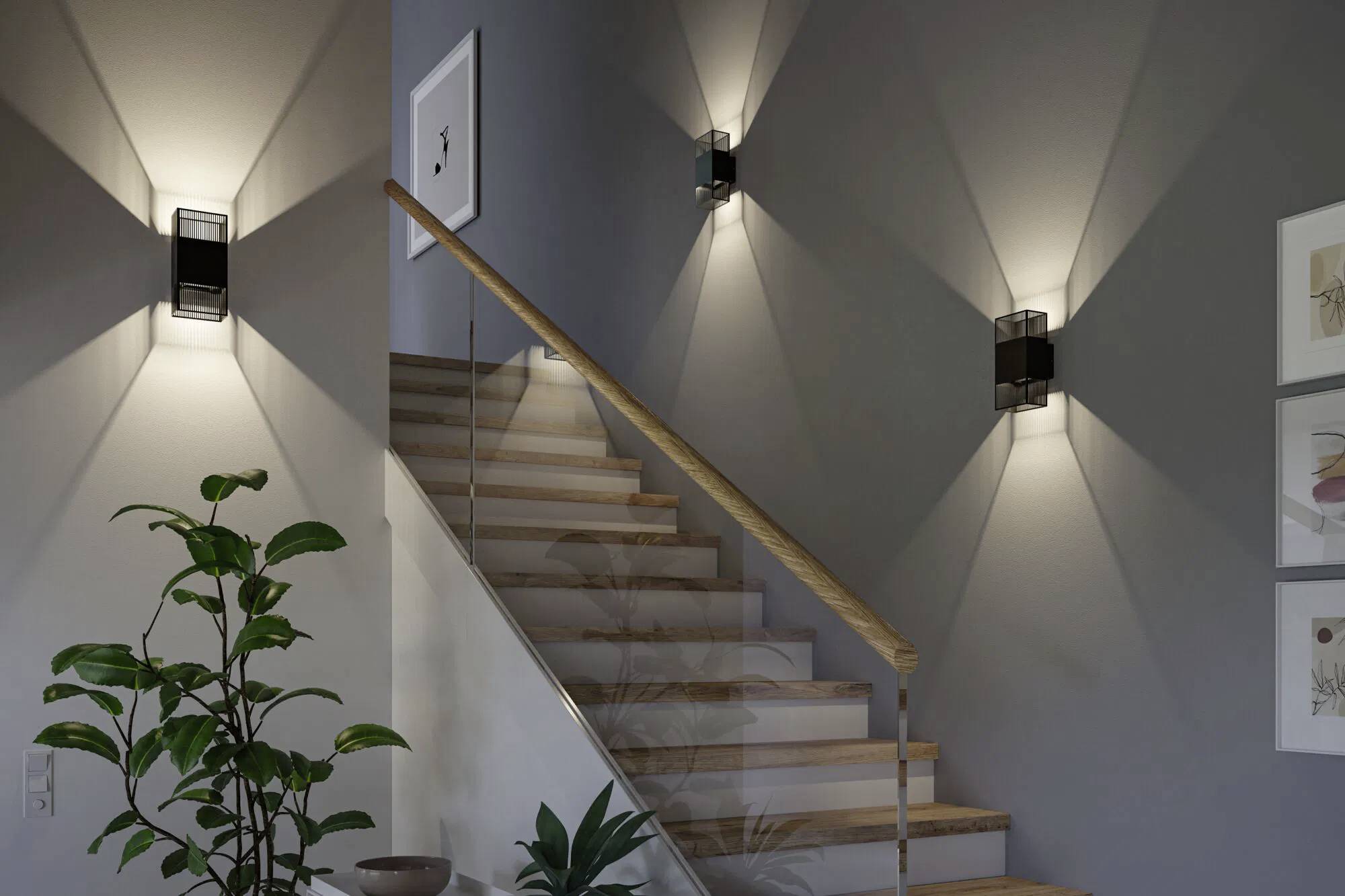 'Een trap met houten treden wordt verlicht door drie moderne wandlampen. Onderaan staat een plant en aan de muur hangt ingelijste kunstwerk.'