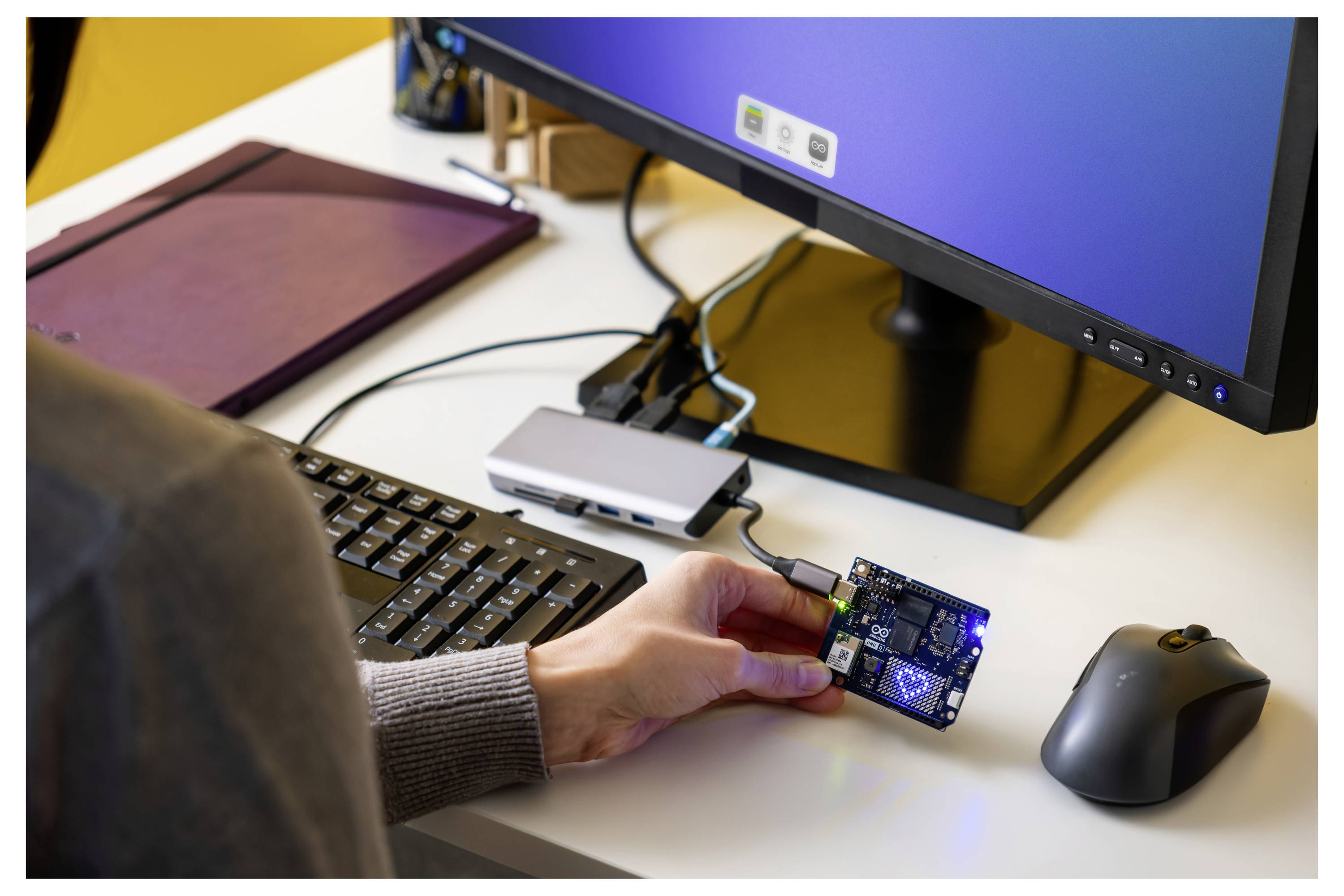 Een persoon houdt een kleine printplaat vast die is aangesloten op een desktopcomputer-installatie, inclusief een monitor, toetsenbord en muis op een bureau.