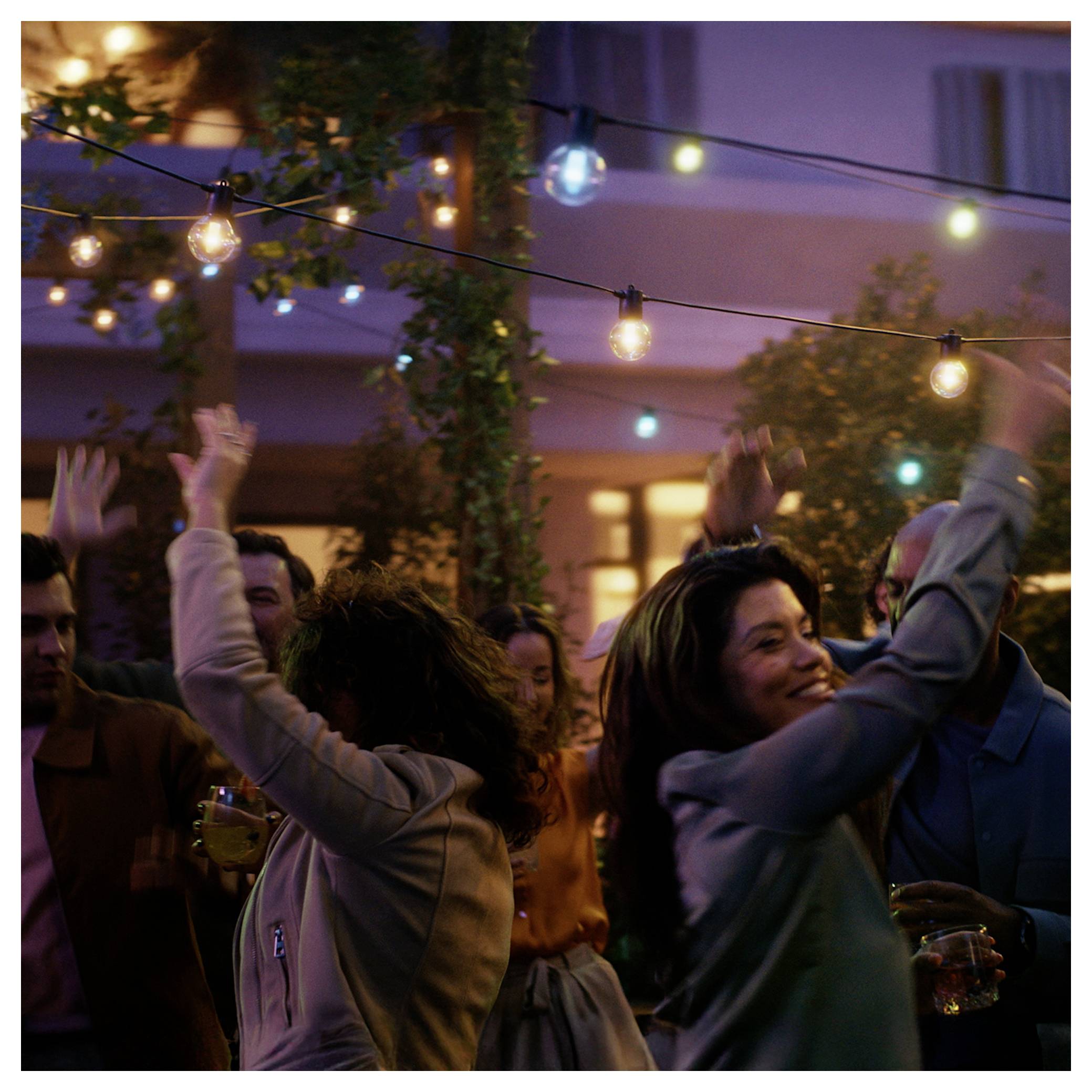 Mensen die vrolijk dansen onder slingerlichten bij een avondelijke buitenfeest, met een feestelijke sfeer en een warmverlicht gebouw op de achtergrond.