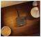 Een draadloze oplader op een houten oppervlak met een verlichte indicator, naast decoratieve objecten zoals een kaars en een kleine witte vaas.