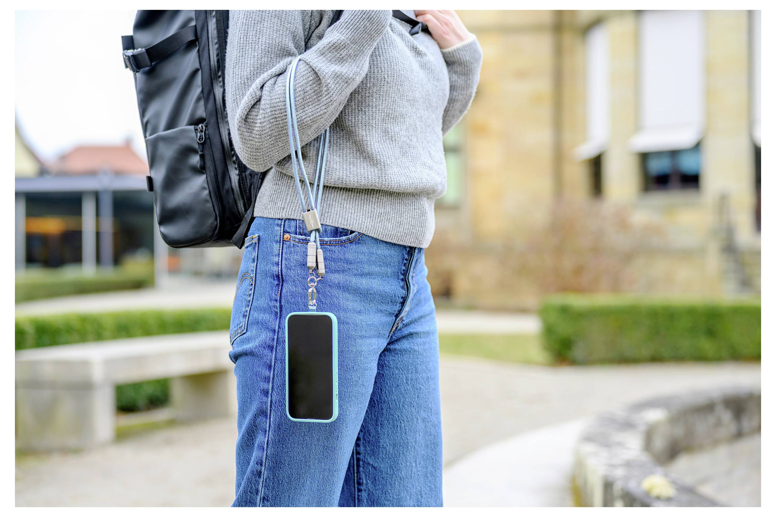 Persoon in casual kleding die een rugzak draagt met een smartphone aan een lanyard vastgemaakt aan de spijkerbroek, met een buitenachtergrond.