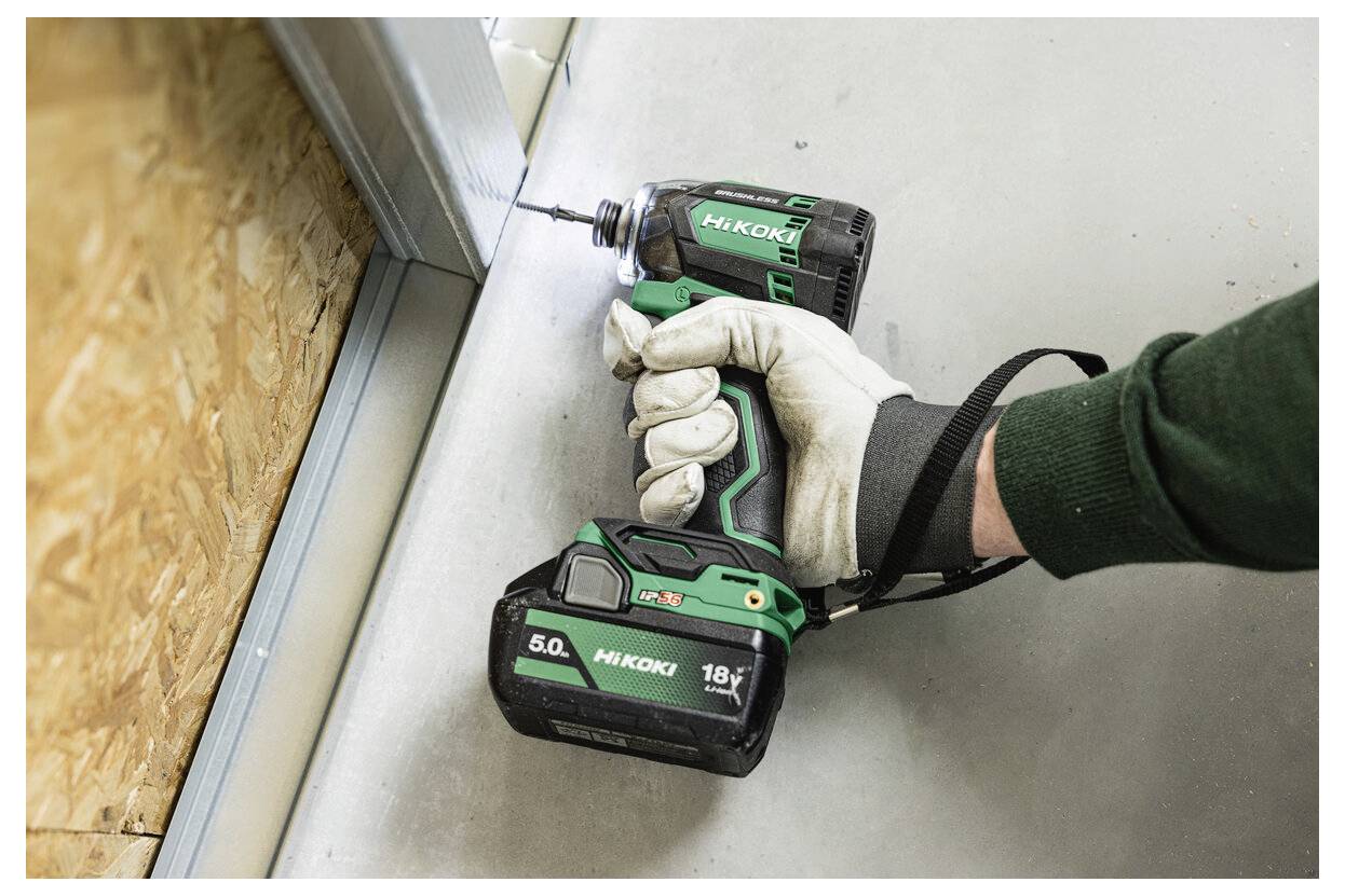 Een persoon die handschoenen draagt, gebruikt een groene accuboormachine om een metalen frame te bevestigen op een betonnen vloer naast een houten muur.