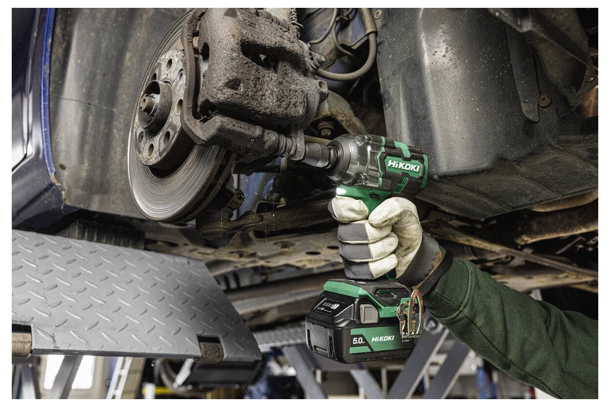 Een gehandschoende hand gebruikt een draadloze slagmoersleutel op de rembehuizing van een auto in een garage. Het gereedschap is groen en zwart, stevig vastgehouden.