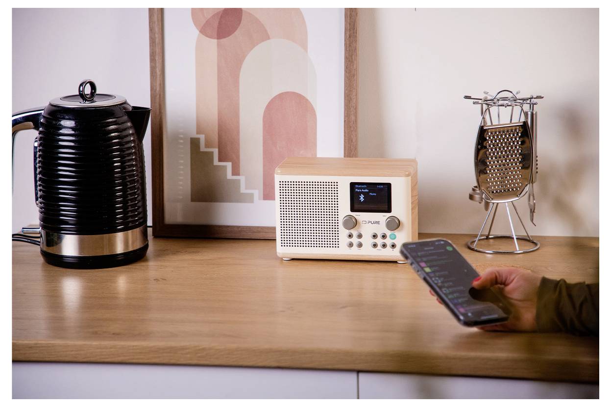 Een aanrecht in de keuken met een waterkoker, ingelijste kunstwerken, een retro radio en een persoon die een smartphone gebruikt.