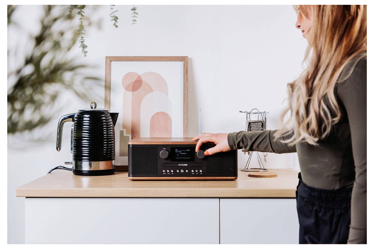 Een persoon draait aan de knop van een radio in retrostijl die staat opgesteld op een houten aanrecht, naast een zwarte waterkoker en abstracte kunst.