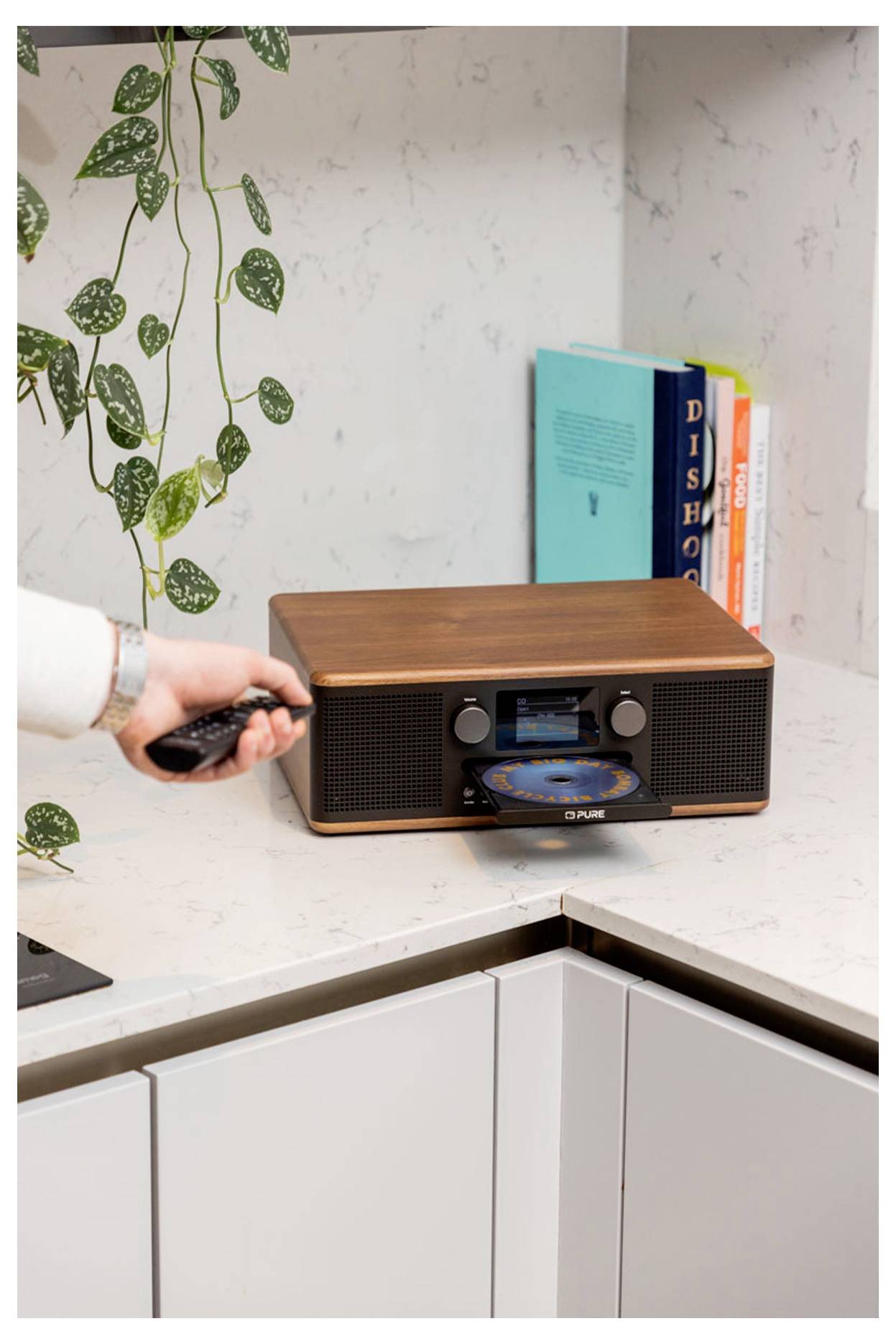 Een persoon bedient een retro-stijl muziekspeler met een cd op een aanrecht; boeken en een plant bevinden zich op de achtergrond.