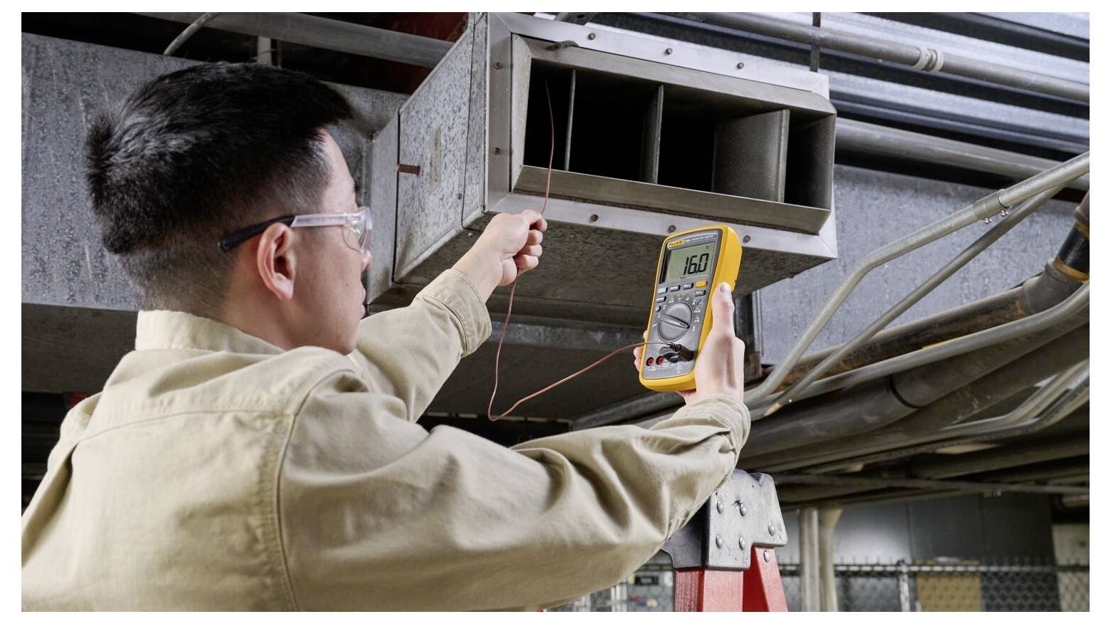 Een persoon die een digitale multimeter gebruikt om spanning te meten nabij een luchtkanaal in een industriële omgeving, met de nadruk op elektrische inspectiewerkzaamheden.
