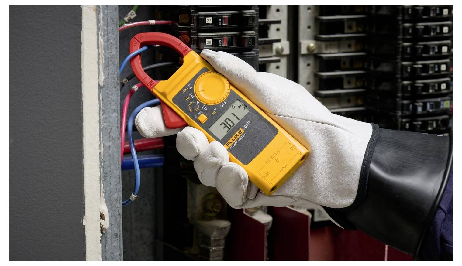 Een persoon met handschoenen gebruikt een gele klemmeterset op elektrische draden in een paneel, waarbij een aflezing van 201,2 wordt weergegeven.