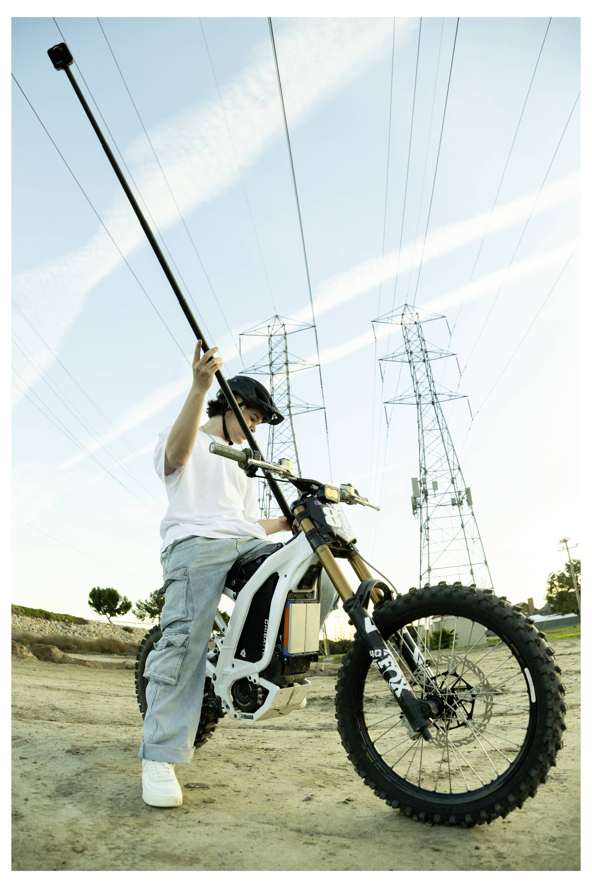 Persoon op een terreinfiets die een lange monopod vasthoudt, met elektriciteitsleidingen en een blauwe lucht op de achtergrond.