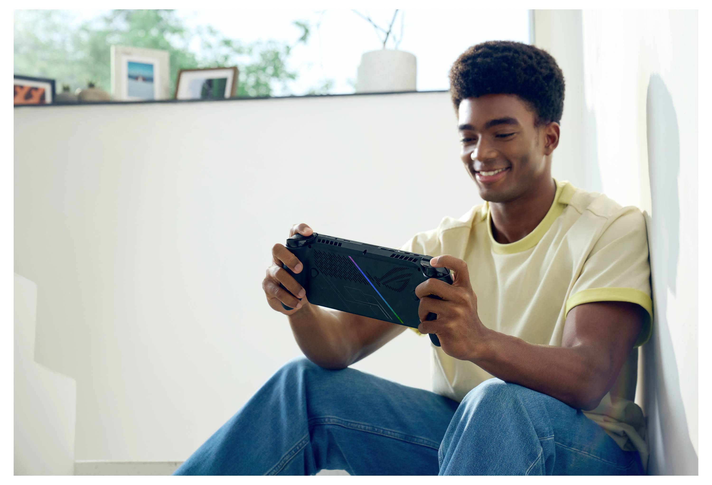 Een persoon die binnen zit, glimlacht terwijl hij een draagbare gameconsole speelt, met ingelijste foto's en een plant zichtbaar op de achtergrond.