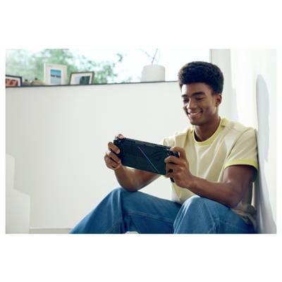 Een persoon die binnen zit, glimlacht terwijl hij een draagbare gameconsole speelt, met ingelijste foto's en een plant zichtbaar op de achtergrond.