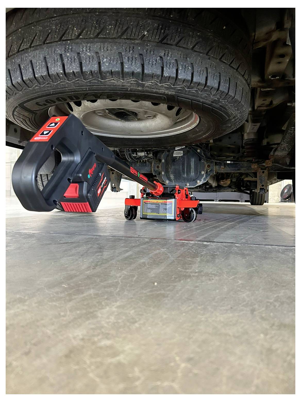 Een autoband die wordt opgetild door een hydraulische krik, waarbij de krikstang en bedieningspaneel zichtbaar zijn op de voorgrond, op een betonnen ondergrond.