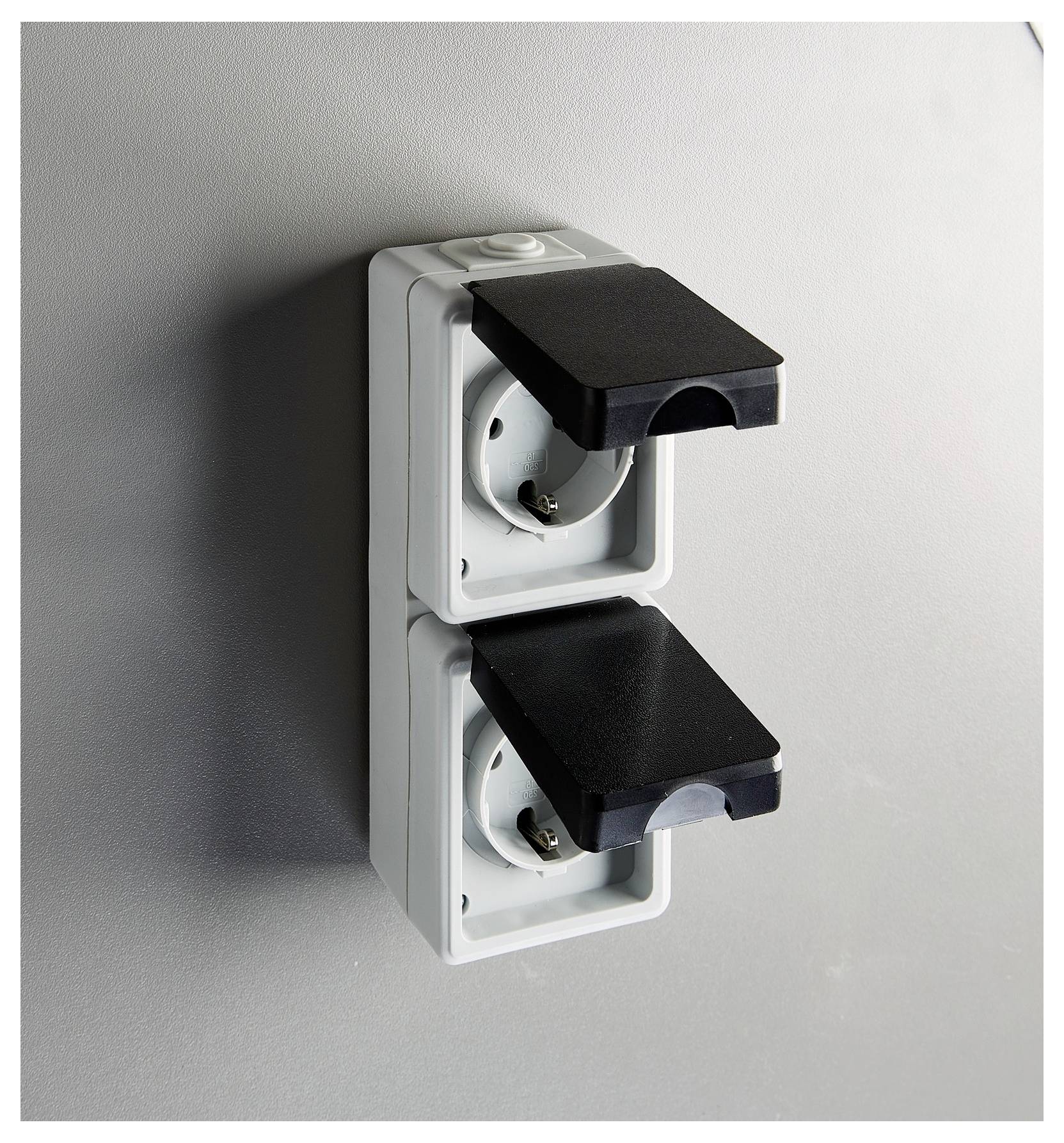 Twee buitenstopcontacten naast elkaar met beschermende zwarte deksels, gemonteerd op een grijze muur, ontworpen voor weerbestendigheid.