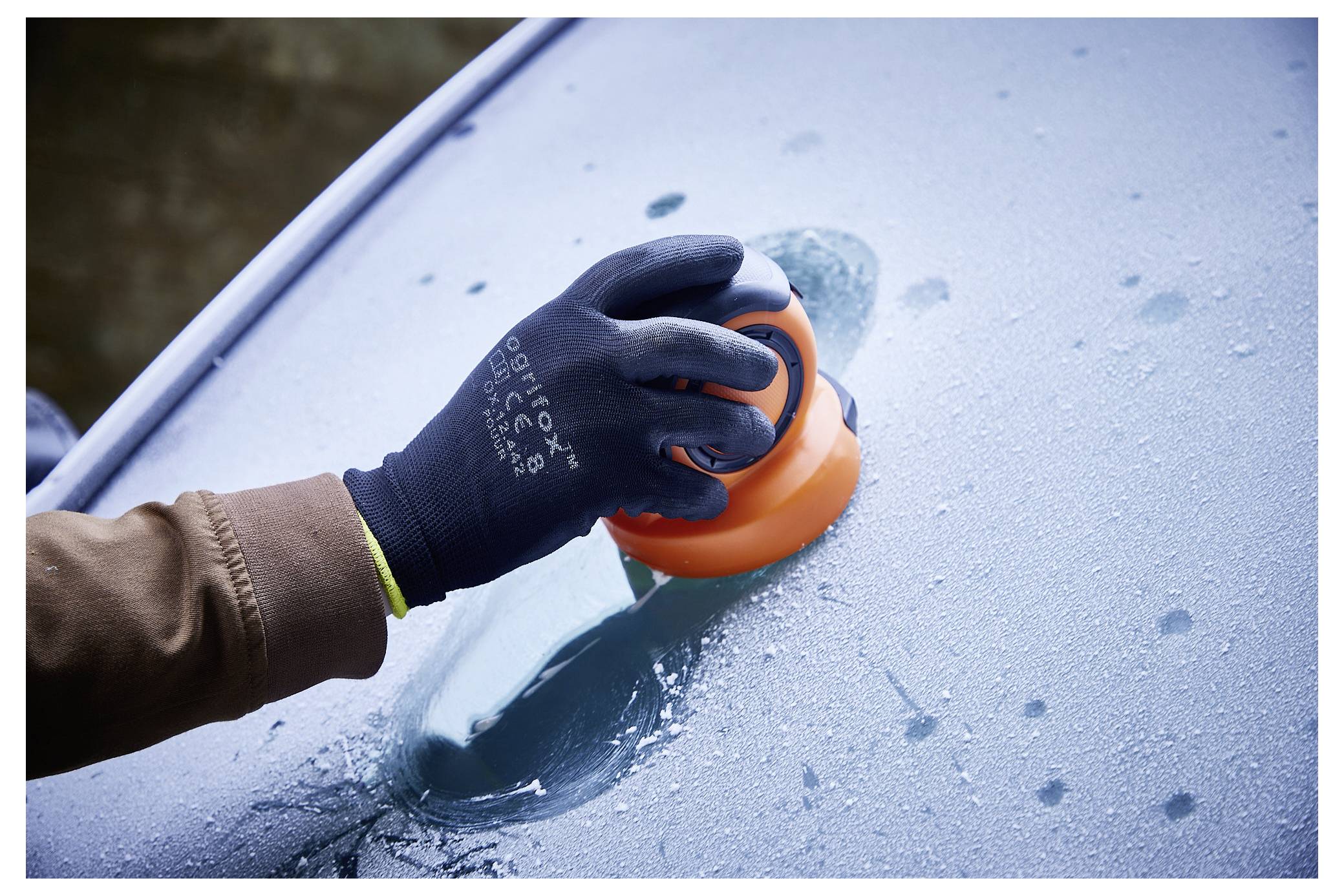 'Een hand met handschoen die een oranje ijskrabber gebruikt om rijp van een autovoorruit schoon te maken.'