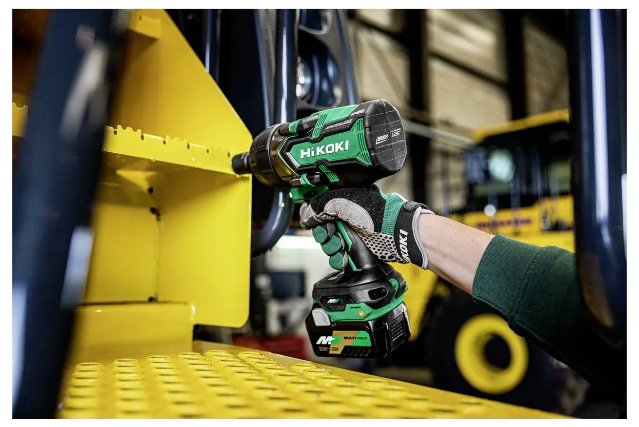 Een hand in een handschoen gebruikt een groene snoerloze slagschroevendraaier om een bout vast te draaien op gele industriële apparatuur in een werkplaatsomgeving.