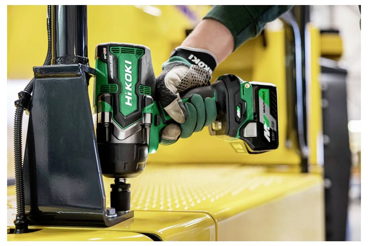 'Een persoon die een groene draadloze accuboor gebruikt op een gele metalen oppervlakte binnenshuis. Op de achtergrond is industriële apparatuur te zien.'