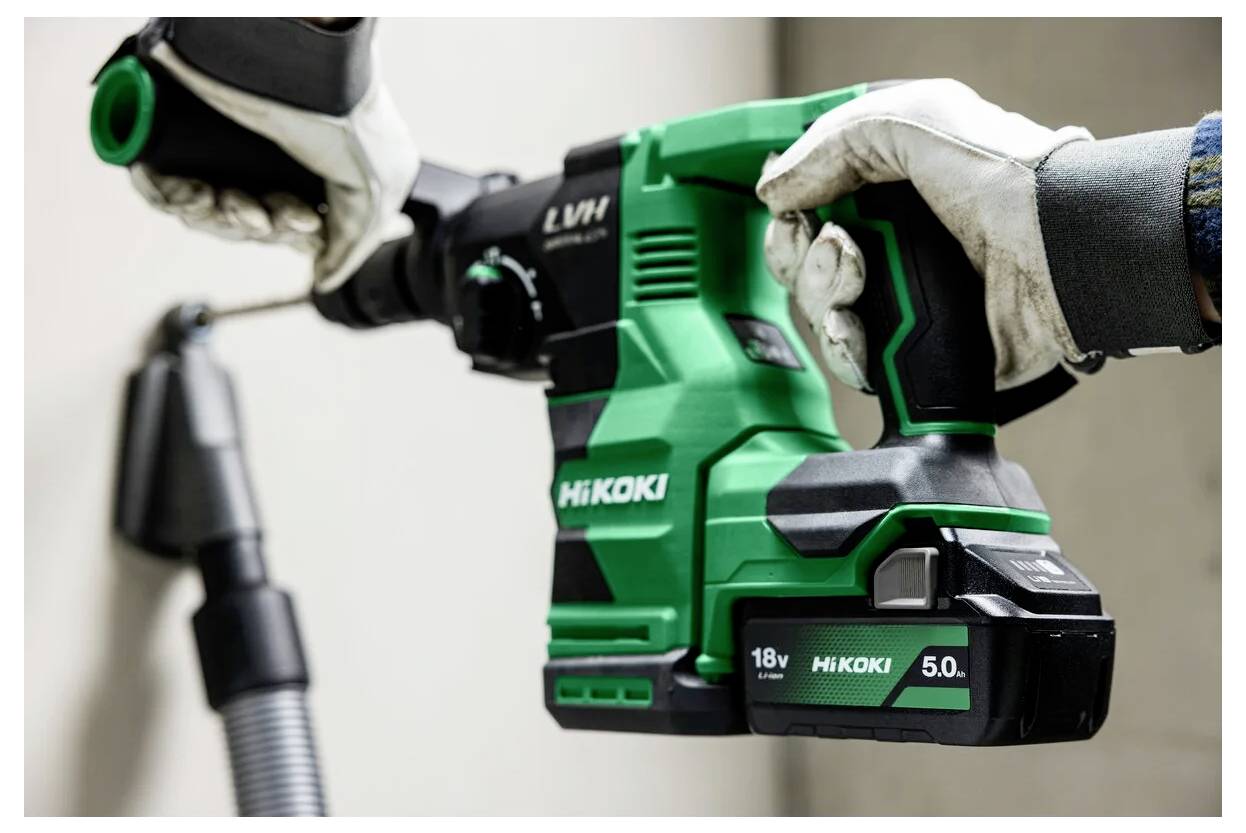 Een persoon met handschoenen gebruikt een groene snoerloze boormachine van het merk 'HiKOKI'. De boormachine heeft een 18V, 5.0Ah accu.