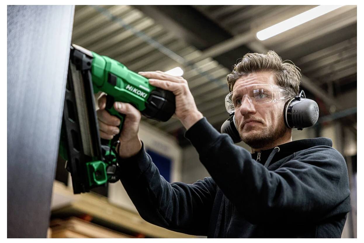 Een persoon die gehoorbescherming en veiligheidsbril draagt, gebruikt een groene spijkerpistool op een houten oppervlak binnen een werkplaats.