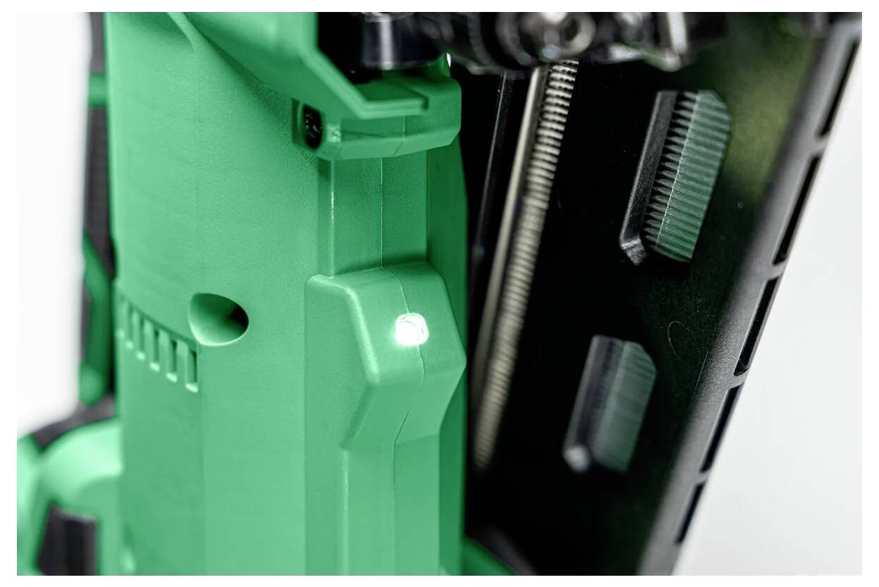 Een close-up van een groen gereedschap, mogelijk een elektrisch gereedschap of nagelpistool, met een zichtbare LED-lamp die aangeeft dat het actief is. Details van metalen onderdelen zijn zichtbaar.