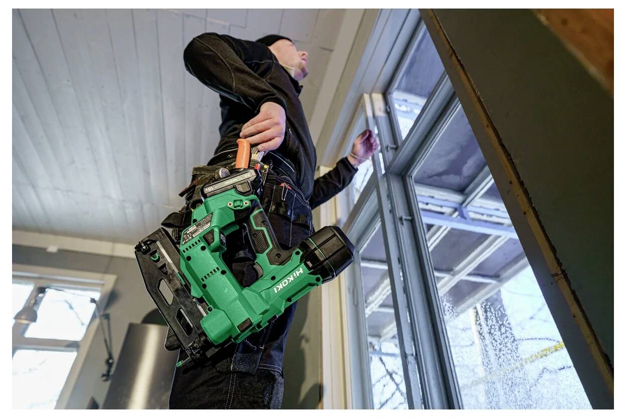 Een persoon in werkkleding staat bij een raam, met een groene accuboormachine in de hand. De omgeving lijkt binnen te zijn, waarschijnlijk bezig met renovatie of inspectie.