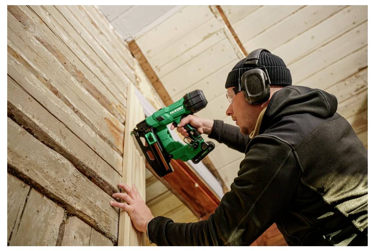 Een persoon met beschermende uitrusting gebruikt een spijkerpistool om houten panelen aan een muur te bevestigen in een kamer die wordt gerenoveerd.