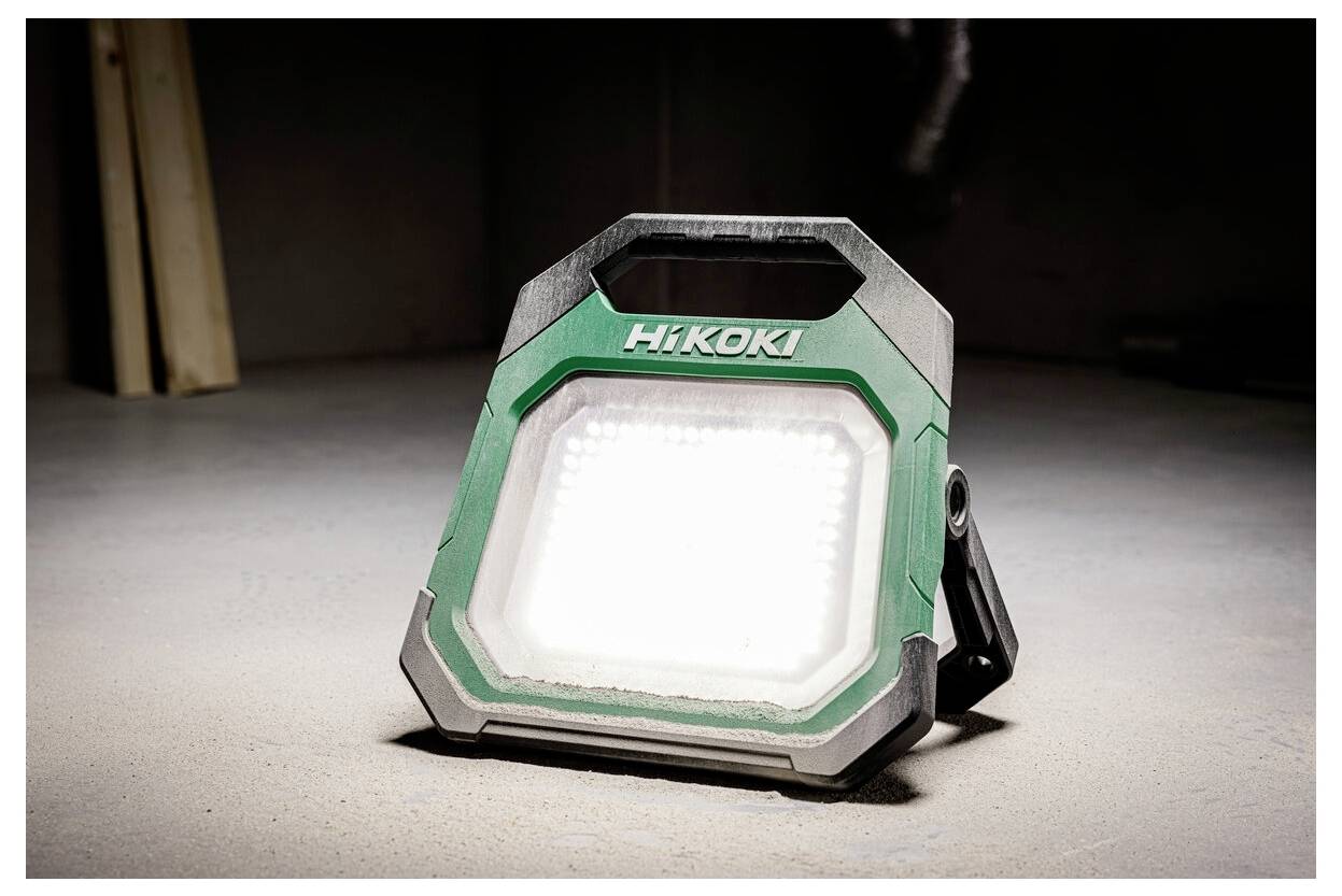 Een draagbare, robuuste LED-werklamp met een groen en zwart frame, gemerkt 'Hikoki', staat op een betonnen oppervlak en verlicht de ruimte.