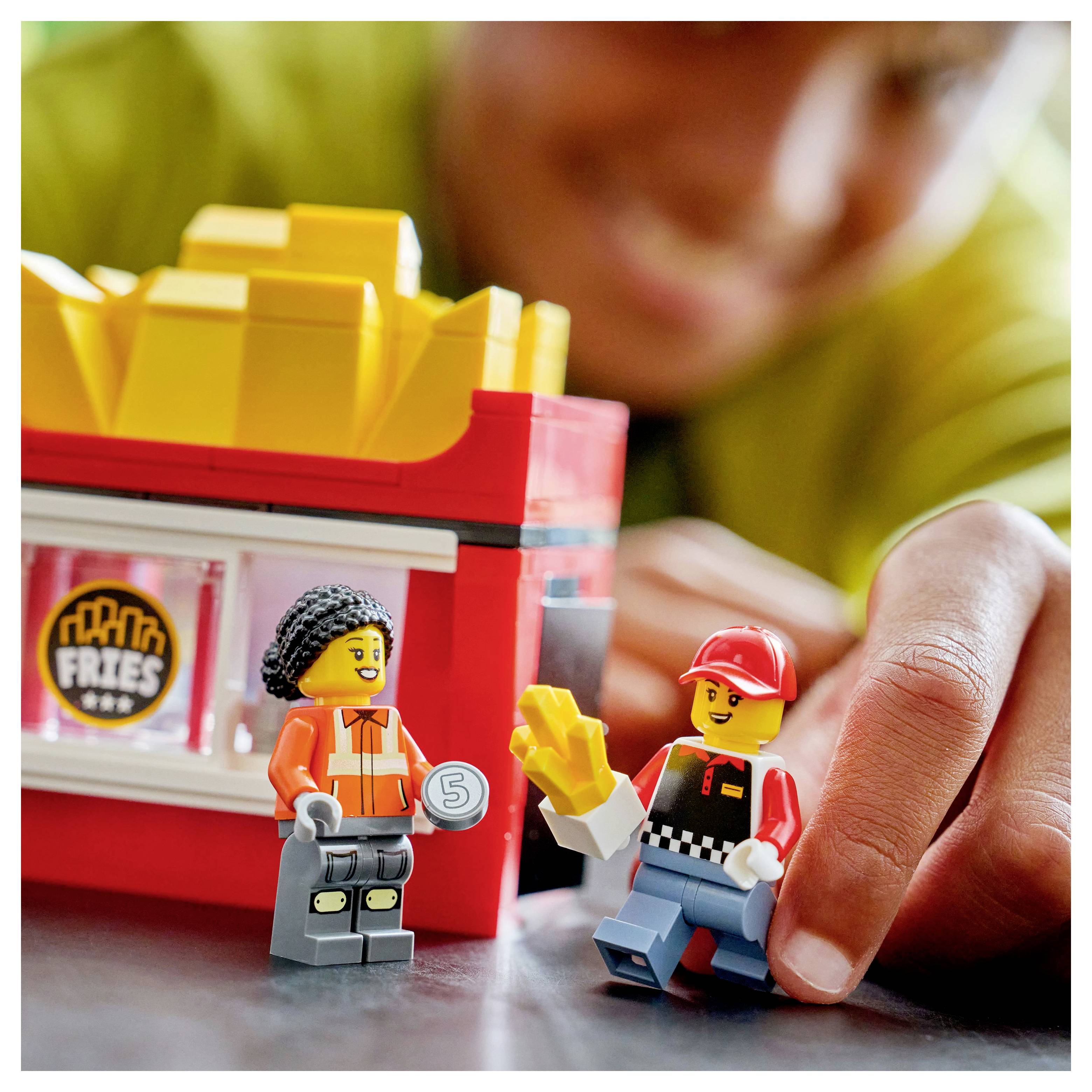 Een kind speelt met speelfiguren die arbeiders in een fastfood-omgeving voorstellen, waarvan één een frietje vasthoudt en een ander wat lijkt op een geldzegel.