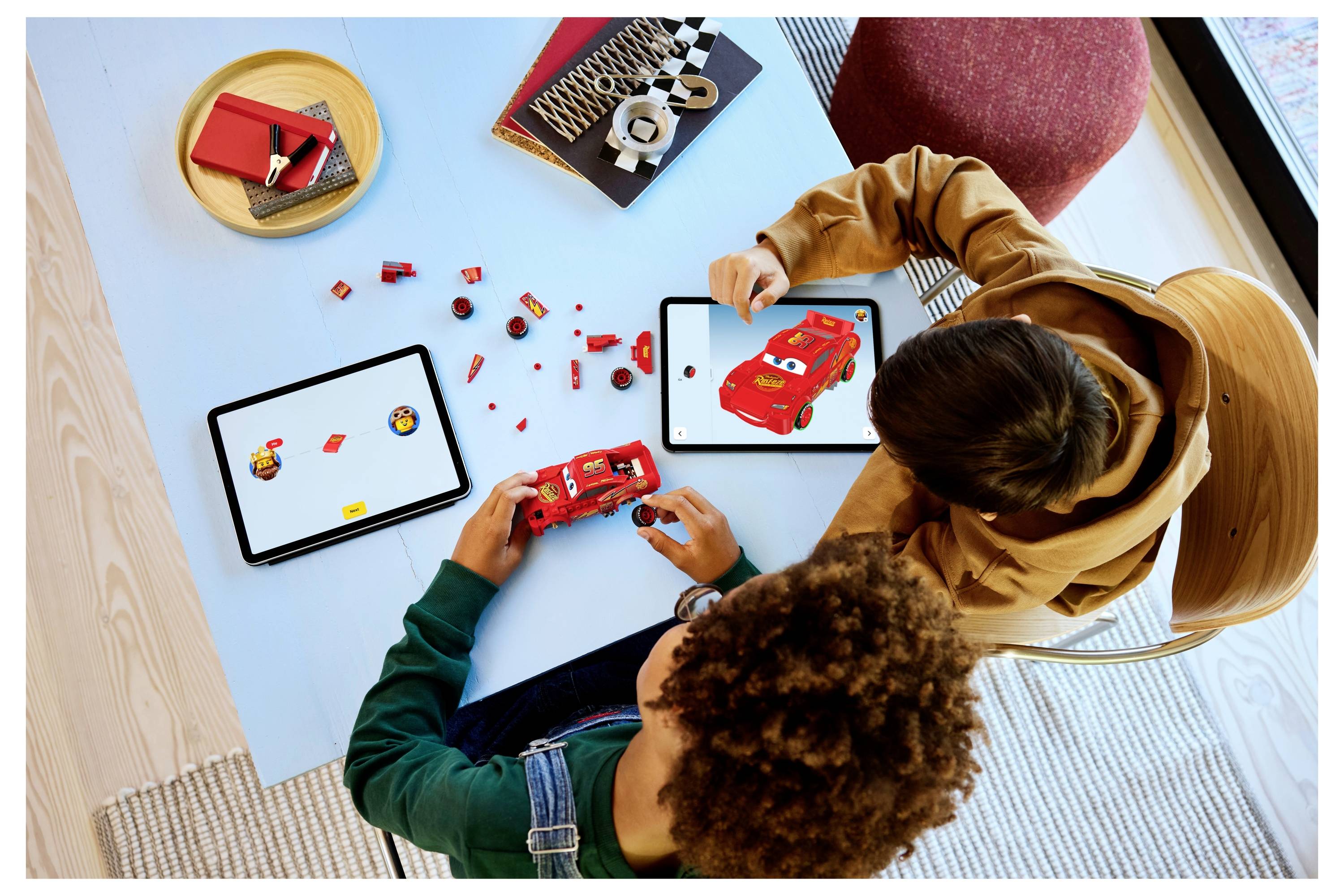 Twee kinderen zitten aan een tafel LEGO-auto's in elkaar te zetten met rode stenen, terwijl ze instructies bekijken op tablets.