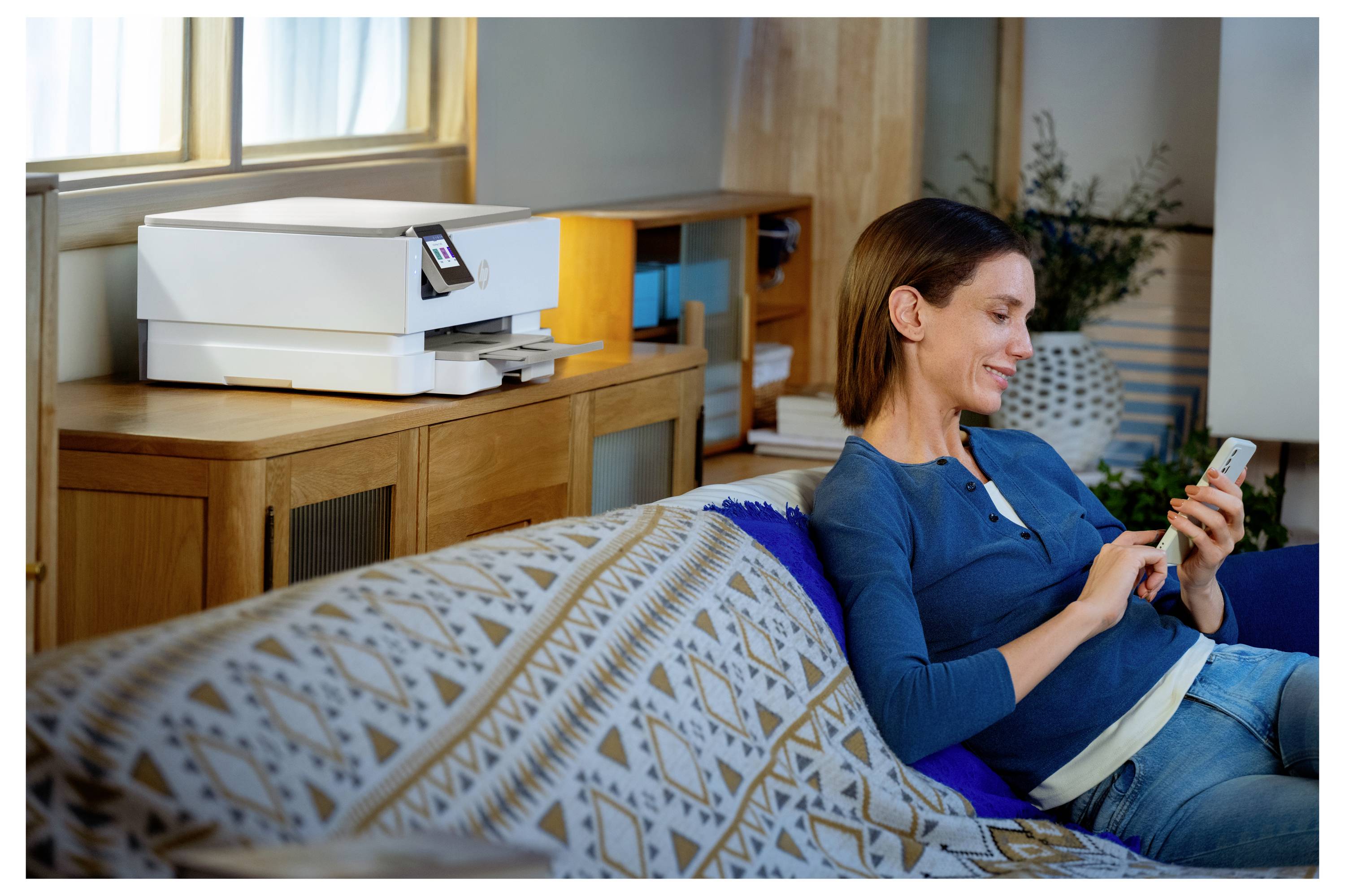 Een vrouw zit op een bank en glimlacht naar haar smartphone. Een moderne printer staat op een plank op de achtergrond, wat een ontspannen thuiskantoor suggereert.