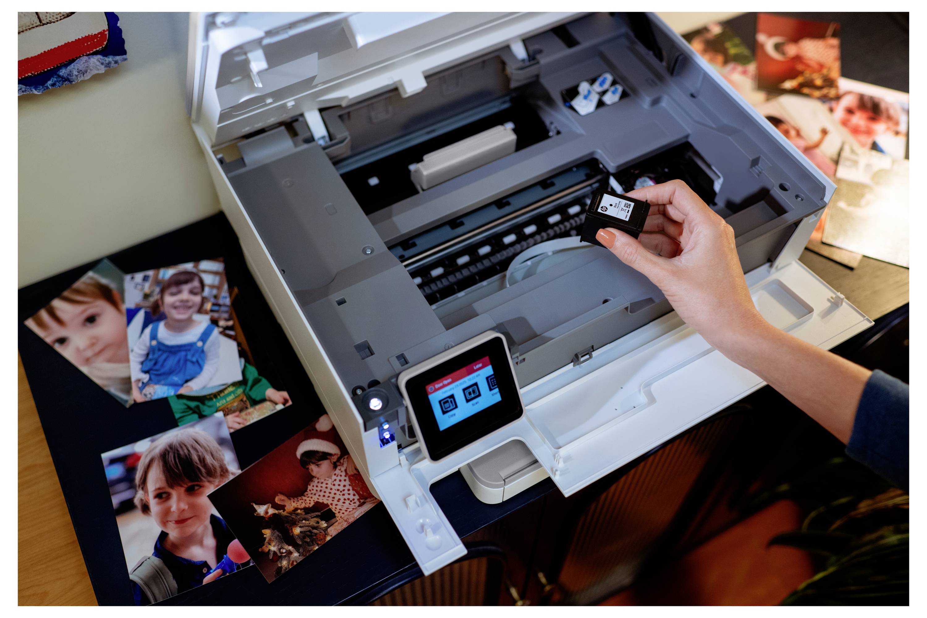 Een persoon vervangt een inktcartridge in een printer, omringd door foto's van kinderen die zijn afgedrukt, wat wijst op een thuisprint-installatie.