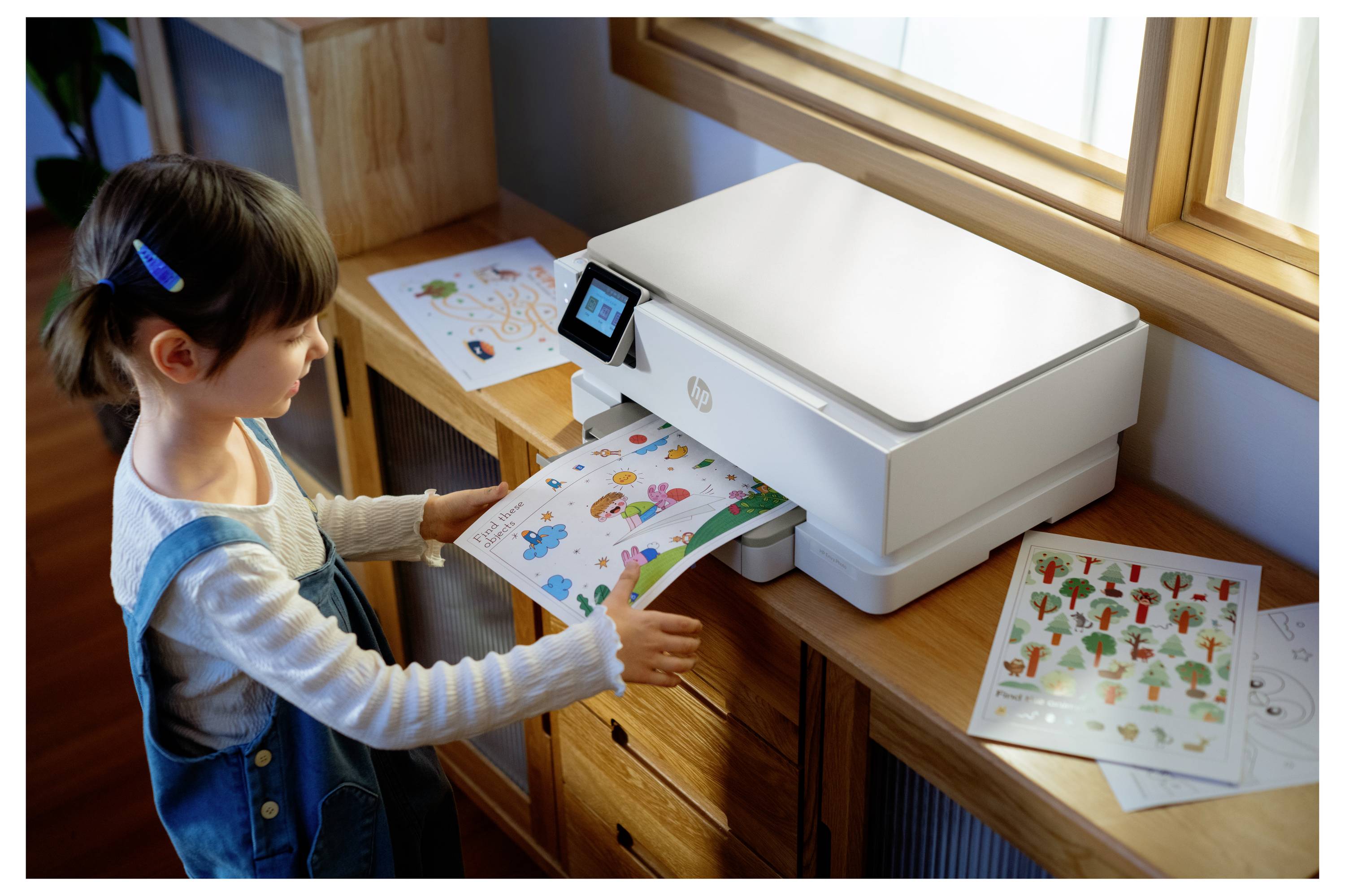 Een kind staat naast een printer en haalt kleurrijke bedrukte vellen met verschillende illustraties op. Kunstbenodigdheden zijn verspreid op de tafel in de buurt.