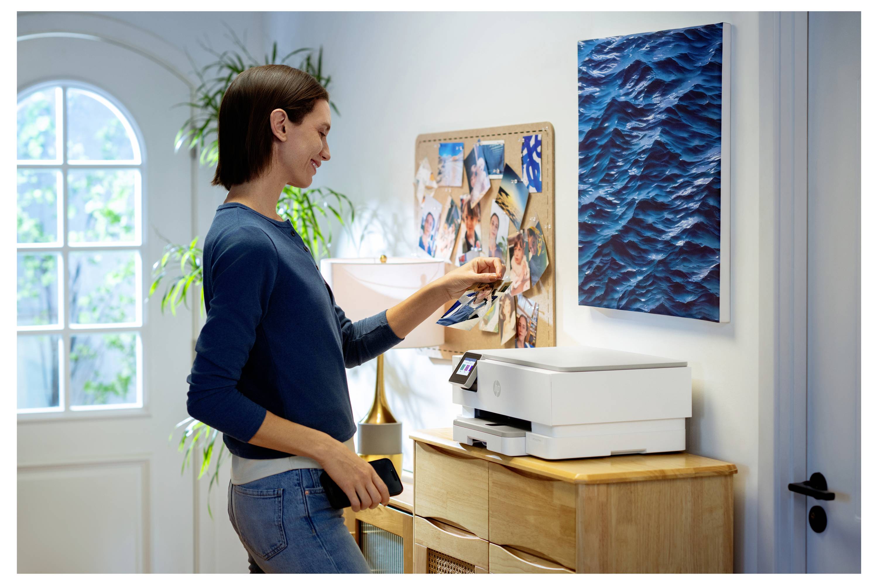Een persoon plaatst een foto van een smartphone op een thuisprinter, staande in een goed verlichte ruimte met een prikbord en een schilderij in de buurt.