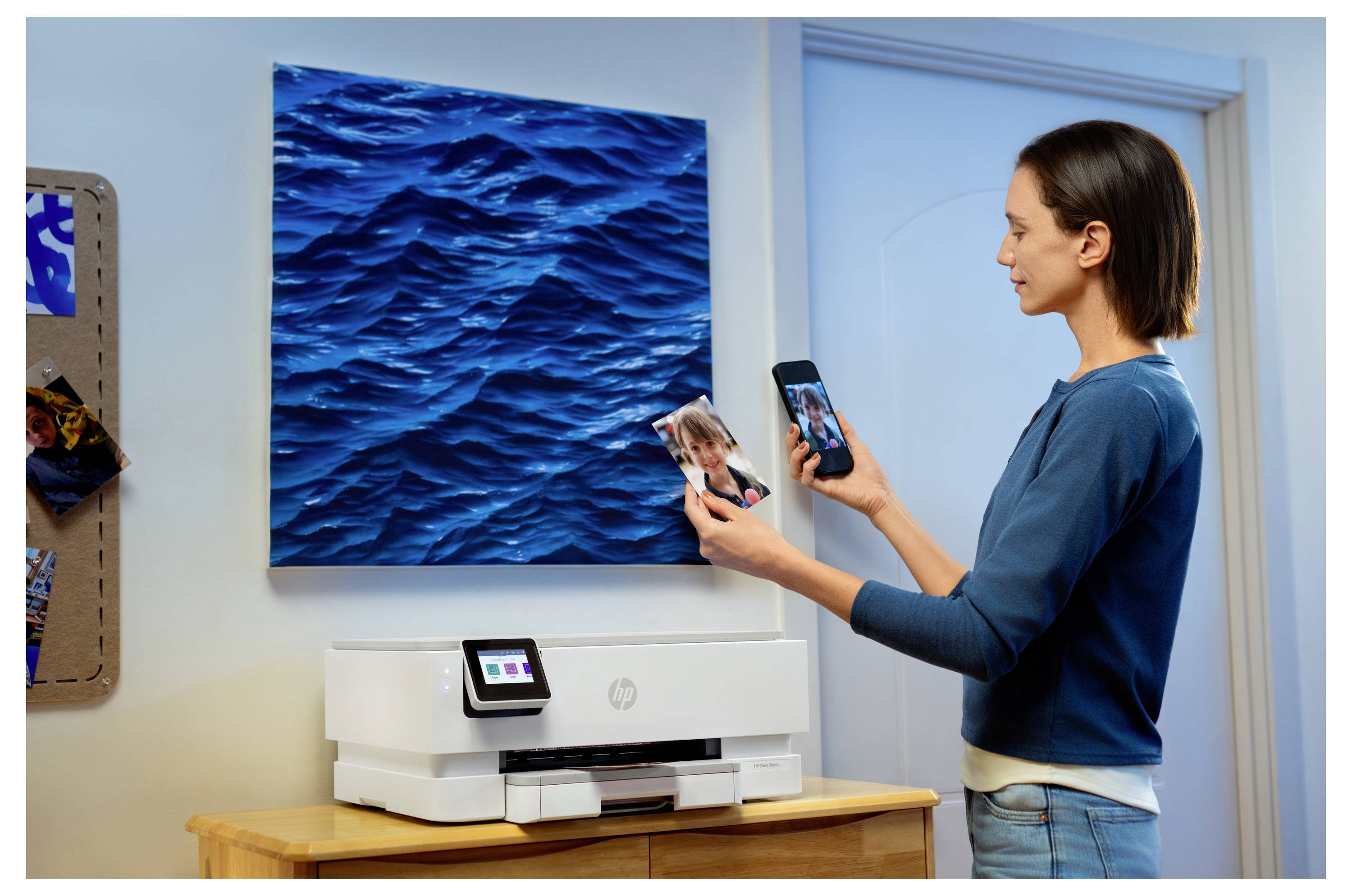 Een vrouw gebruikt een smartphone naast een printer, terwijl ze een afgedrukte foto van een kind vasthoudt. Een blauw oceaankunstwerk hangt aan de muur boven de printer.